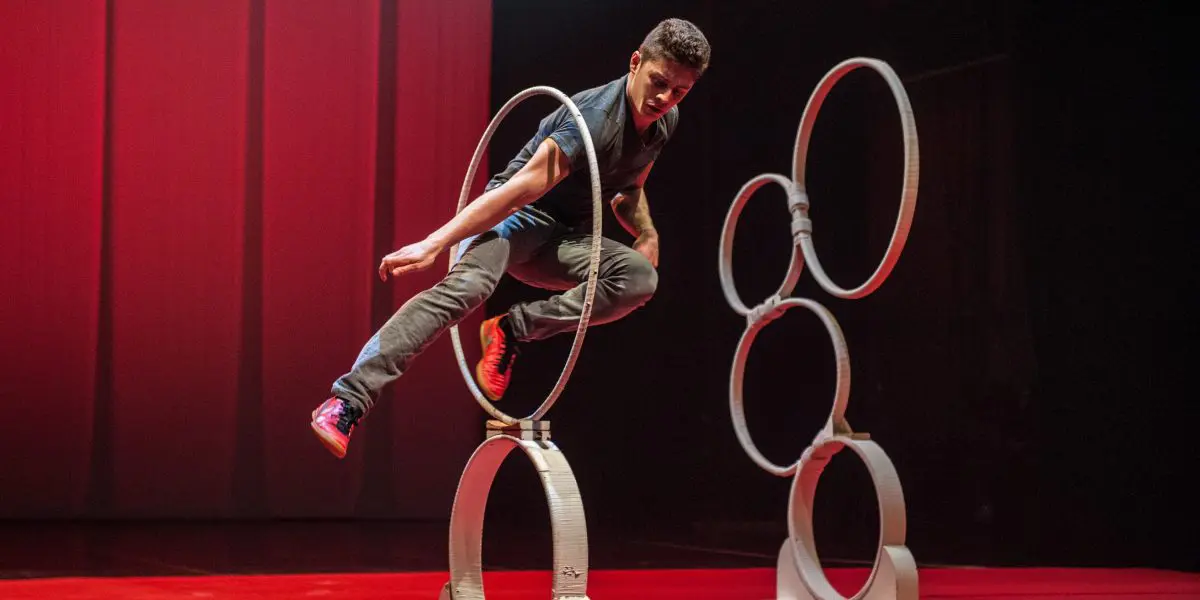 Un homme en tenue décontractée saute habilement à travers un anneau vertical sur scène, évoquant l'agilité de Quentin Gréco, avec d'autres anneaux empilés en arrière-plan et un éclairage rouge dramatique.