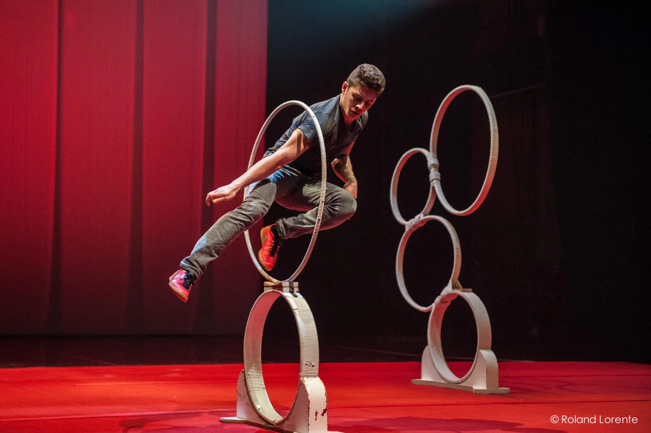 Un homme en tenue décontractée saute habilement à travers un anneau vertical sur scène, évoquant l'agilité de Quentin Gréco, avec d'autres anneaux empilés en arrière-plan et un éclairage rouge dramatique.