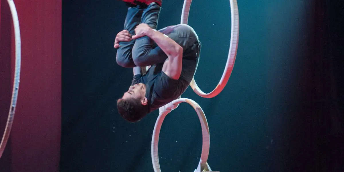 Un artiste en plein vol fait un saut périlleux arrière à travers des cerceaux empilés sur une scène au sol rouge, à l'éclairage dramatique et à l'arrière-plan sombre, rappelant les numéros audacieux de Quentin Gréco.