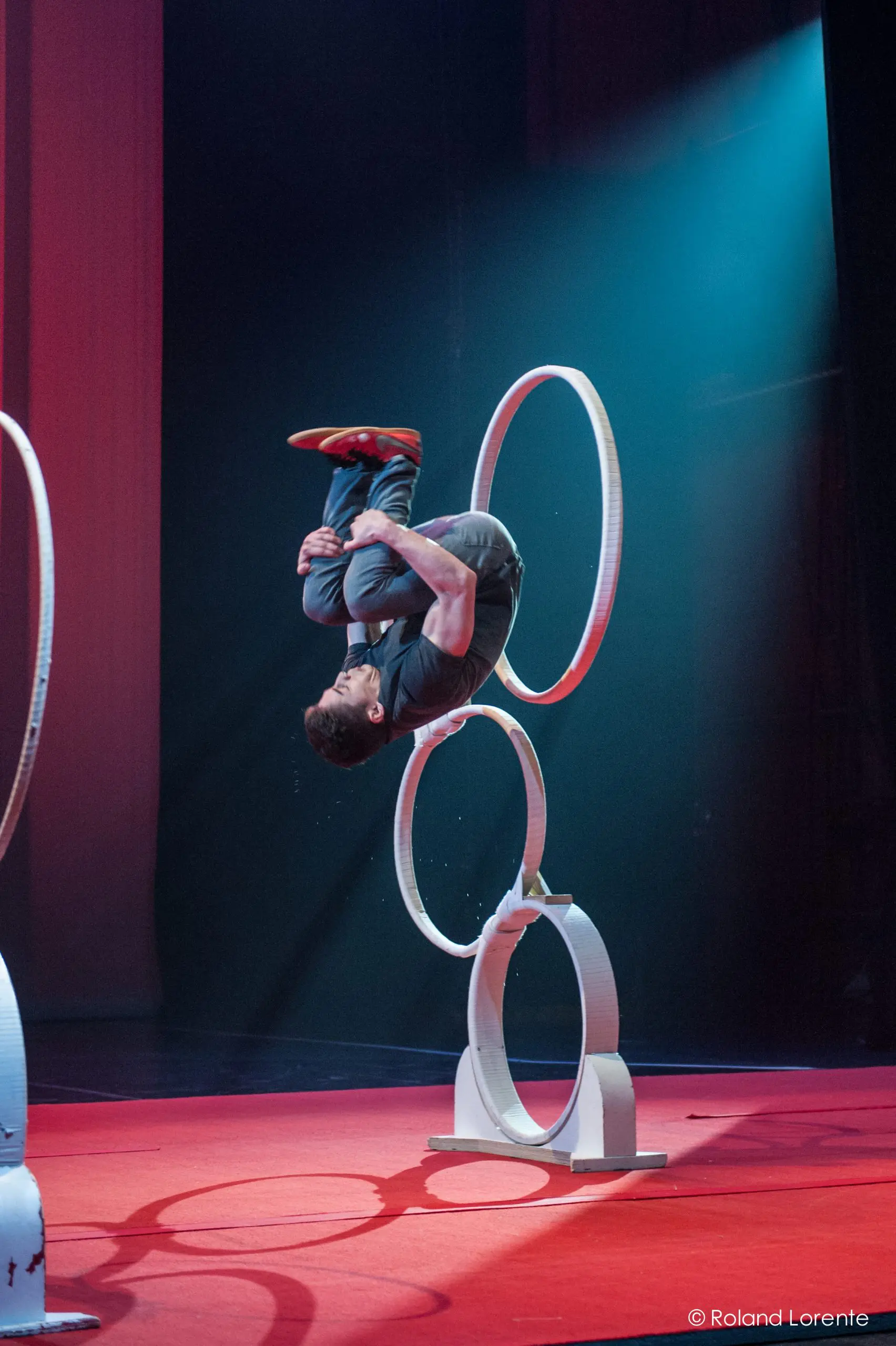 Un artiste en plein vol fait un saut périlleux arrière à travers des cerceaux empilés sur une scène au sol rouge, à l'éclairage dramatique et à l'arrière-plan sombre, rappelant les numéros audacieux de Quentin Gréco.