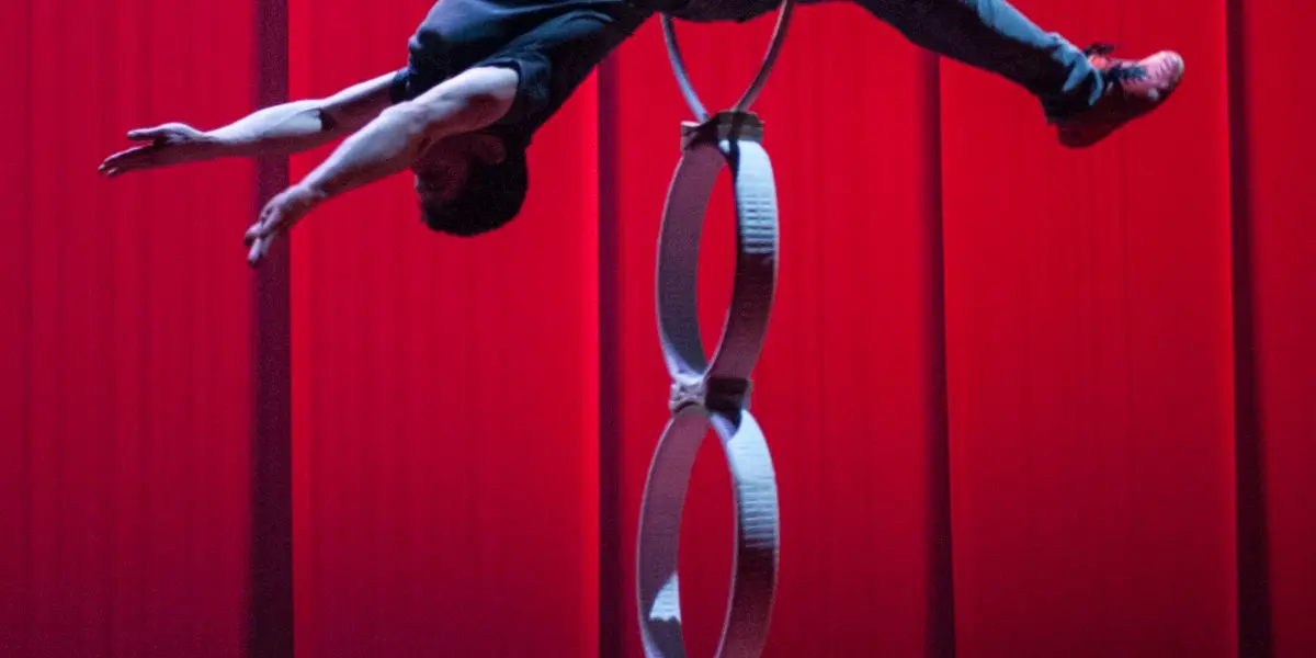 Un artiste, qui rappelle Quentin Gréco ou Evan Tomlinson Weintraub, se tient en équilibre horizontal, les bras tendus, sur une pile de quatre anneaux, soutenue par sa taille, sur un fond de scène rouge.