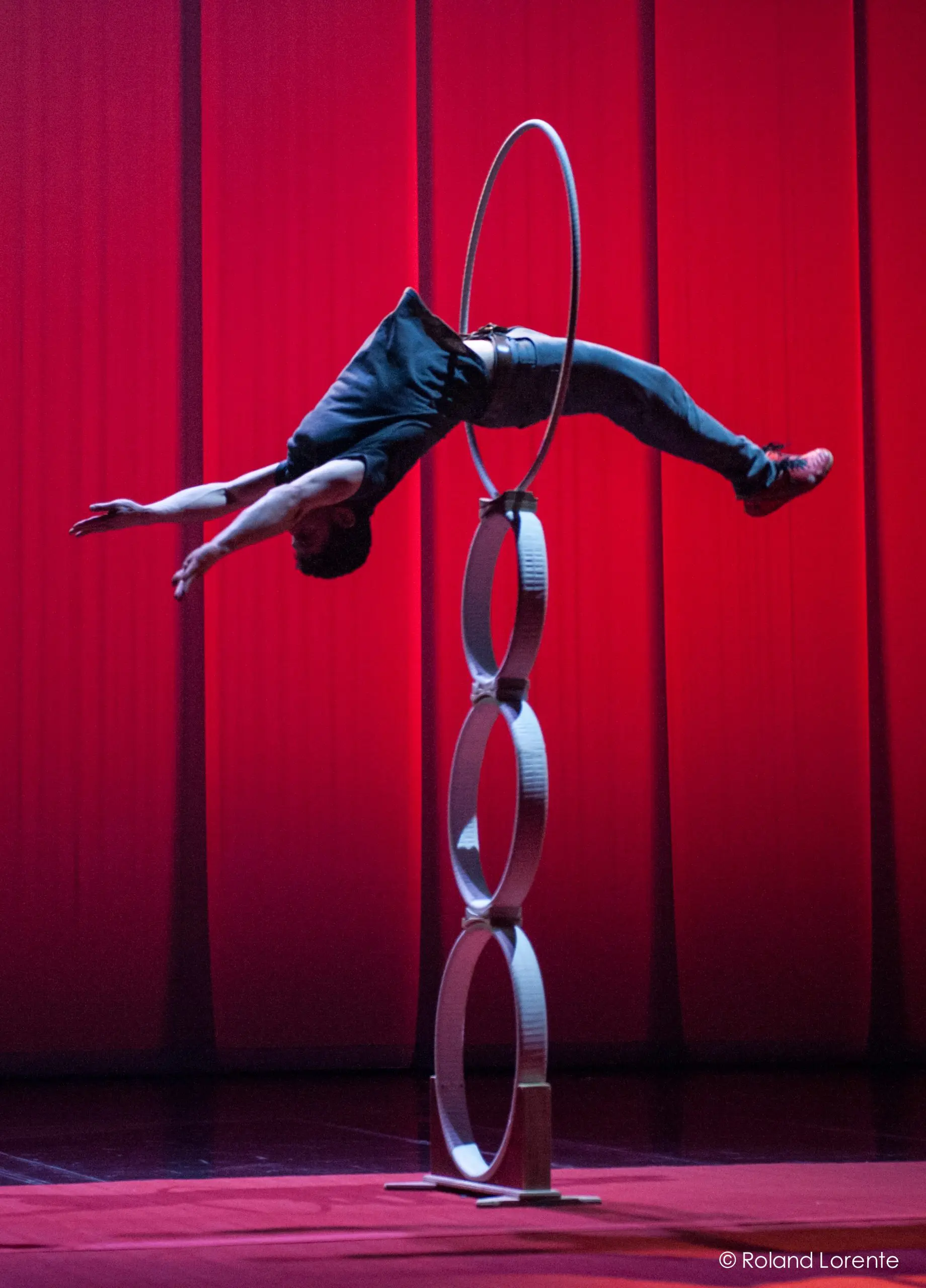 Un artiste, qui rappelle Quentin Gréco ou Evan Tomlinson Weintraub, se tient en équilibre horizontal, les bras tendus, sur une pile de quatre anneaux, soutenue par sa taille, sur un fond de scène rouge.