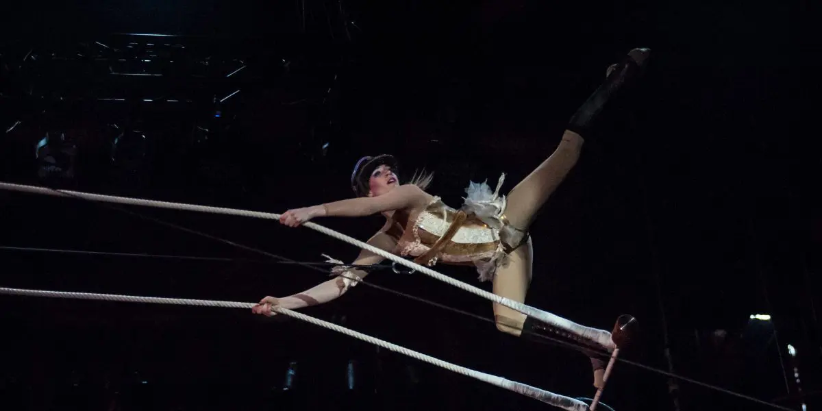 L'artiste de cirque Roxane Gilliand, vêtue d'un costume et d'un chapeau dorés, se tient gracieusement en équilibre sur des cordes raides - une main agrippée à la corde, une jambe tendue vers le haut - tandis que l'éclairage dramatique de la scène met en valeur son numéro époustouflant.