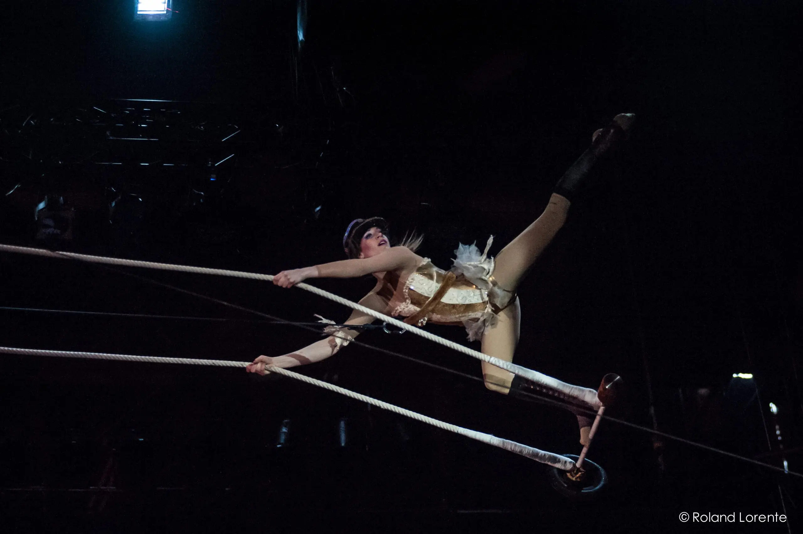 L'artiste de cirque Roxane Gilliand, vêtue d'un costume et d'un chapeau dorés, se tient gracieusement en équilibre sur des cordes raides - une main agrippée à la corde, une jambe tendue vers le haut - tandis que l'éclairage dramatique de la scène met en valeur son numéro époustouflant.