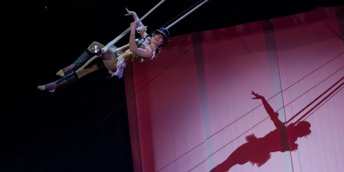 Une trapéziste vêtue d'un costume doré évolue dans les airs, son ombre se projetant de manière spectaculaire sur une toile de fond rose - un spectacle époustouflant de Roxane Gilliand.