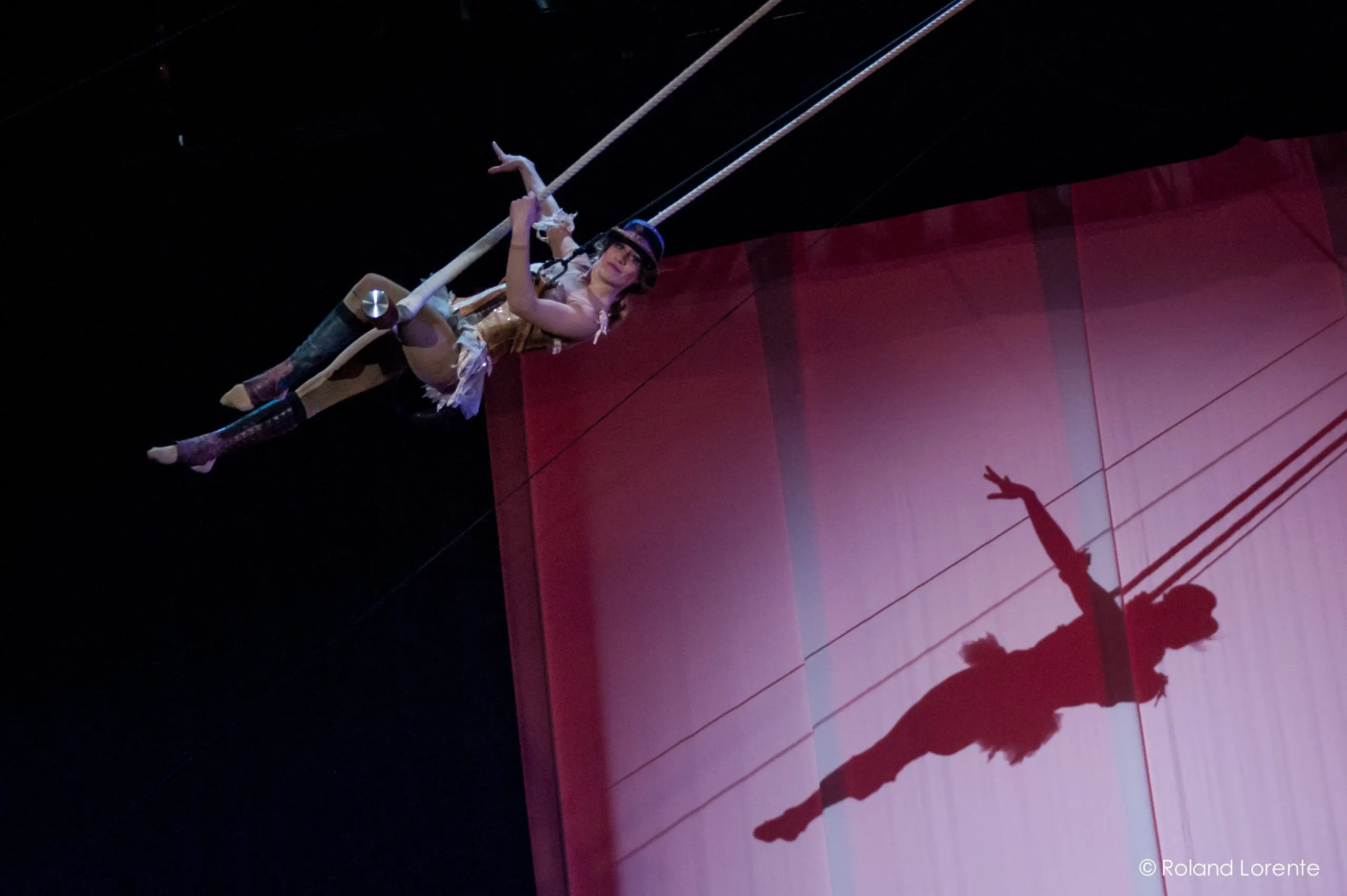 Une trapéziste vêtue d'un costume doré évolue dans les airs, son ombre se projetant de manière spectaculaire sur une toile de fond rose - un spectacle époustouflant de Roxane Gilliand.