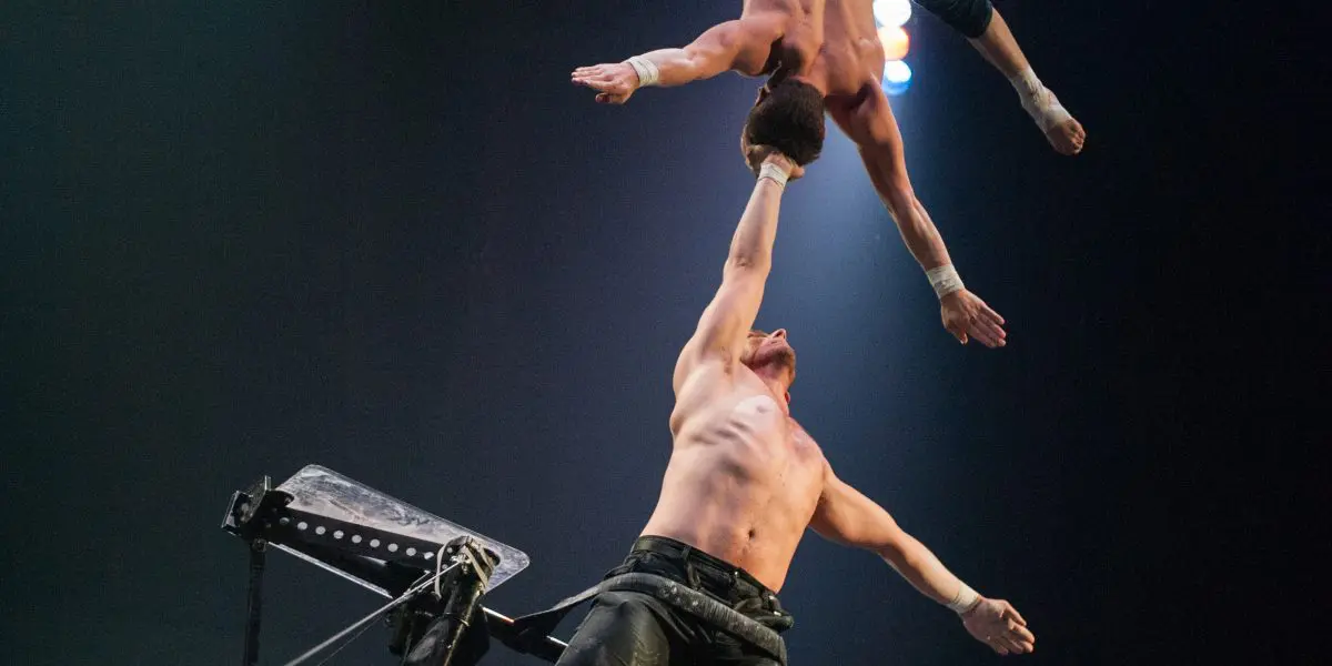 Maxime Blanckaert et Nathan Briscoe réalisent un numéro d'équilibre époustouflant en hauteur ; l'un se tient sur une plate-forme, l'autre en équilibre sur un bras, tandis que des éclairages spectaculaires mettent en valeur leur force et leur habileté.