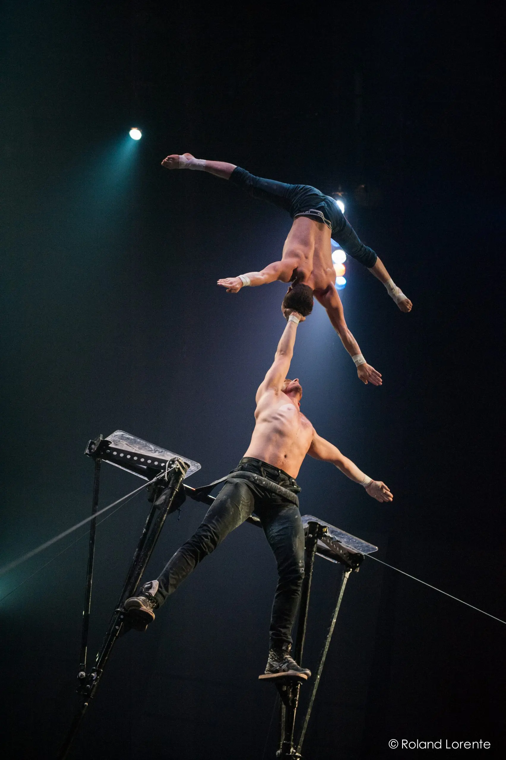 Maxime Blanckaert et Nathan Briscoe réalisent un numéro d'équilibre époustouflant en hauteur ; l'un se tient sur une plate-forme, l'autre en équilibre sur un bras, tandis que des éclairages spectaculaires mettent en valeur leur force et leur habileté.