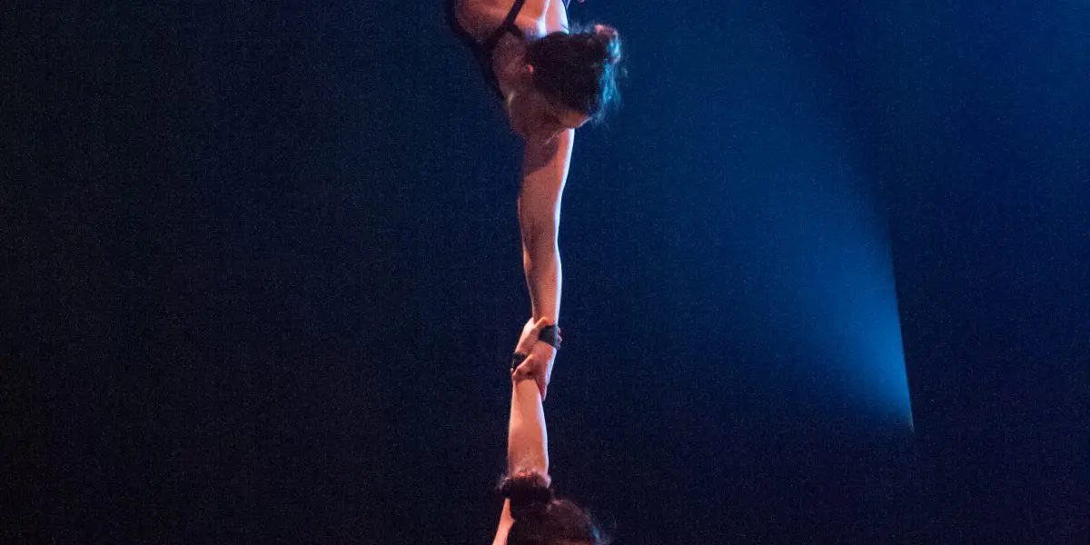 Deux acrobates féminines évoluent sur un trapèze ; l'une est suspendue à l'envers, tenant le bras de l'autre qui est assise en dessous, toutes deux en costumes sombres sur une toile de fond dramatique, incarnant l'élégance et la force de Virginie Gerbeau.