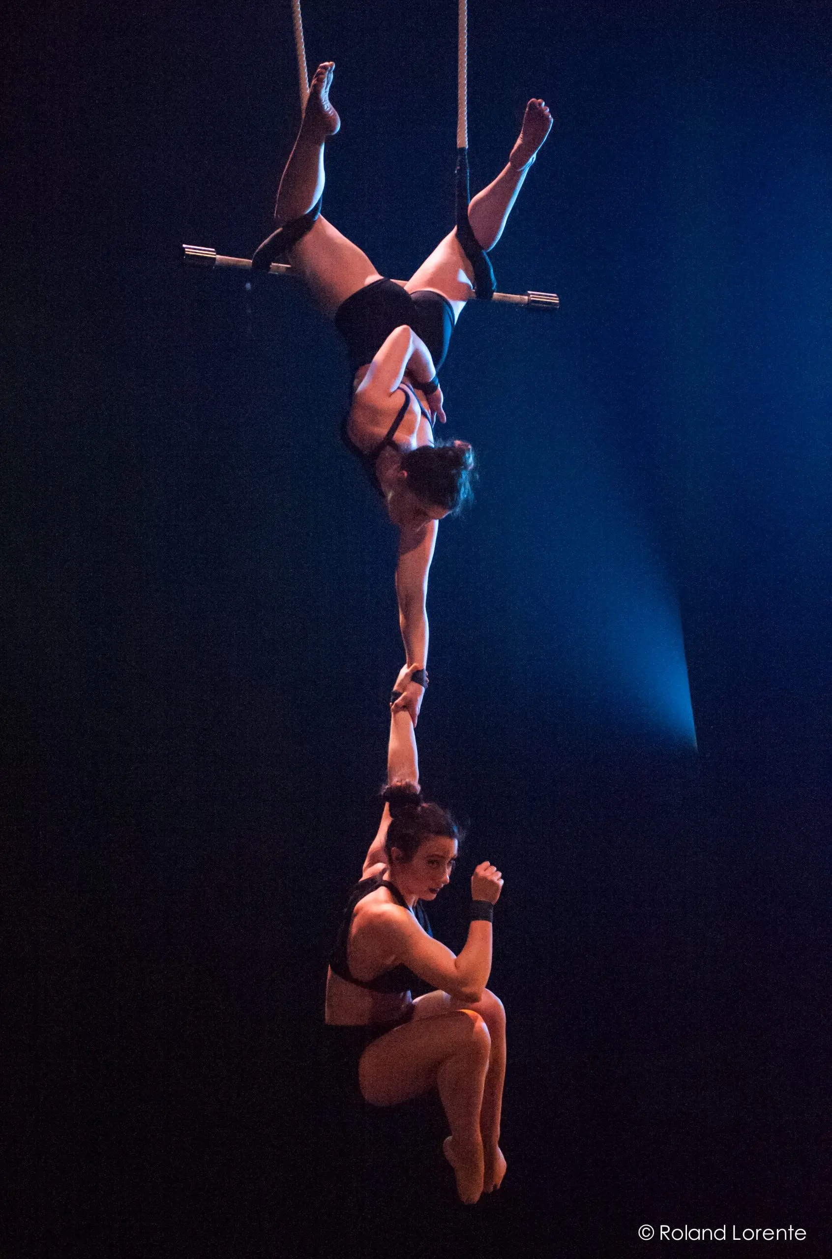Deux acrobates féminines évoluent sur un trapèze ; l'une est suspendue à l'envers, tenant le bras de l'autre qui est assise en dessous, toutes deux en costumes sombres sur une toile de fond dramatique, incarnant l'élégance et la force de Virginie Gerbeau.