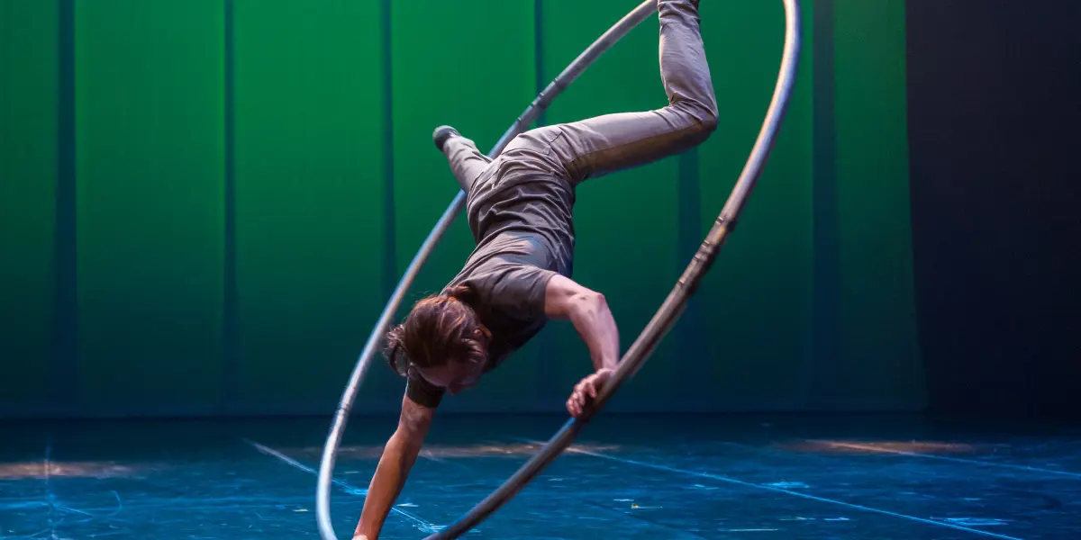 Cory Marsh se tient en équilibre à l'envers dans un grand cerceau métallique, se soutenant d'une main sur une scène éclairée par des lumières bleues et vertes.