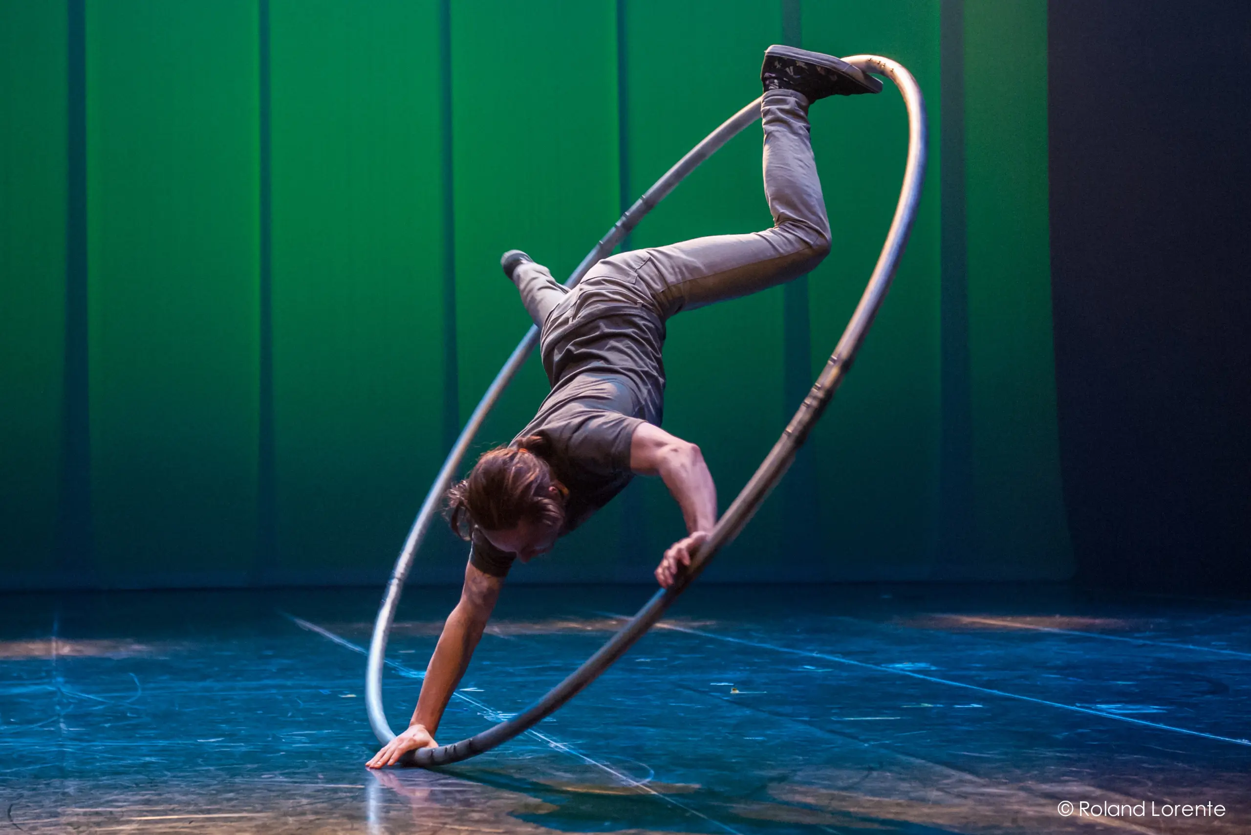 Cory Marsh se tient en équilibre à l'envers dans un grand cerceau métallique, se soutenant d'une main sur une scène éclairée par des lumières bleues et vertes.