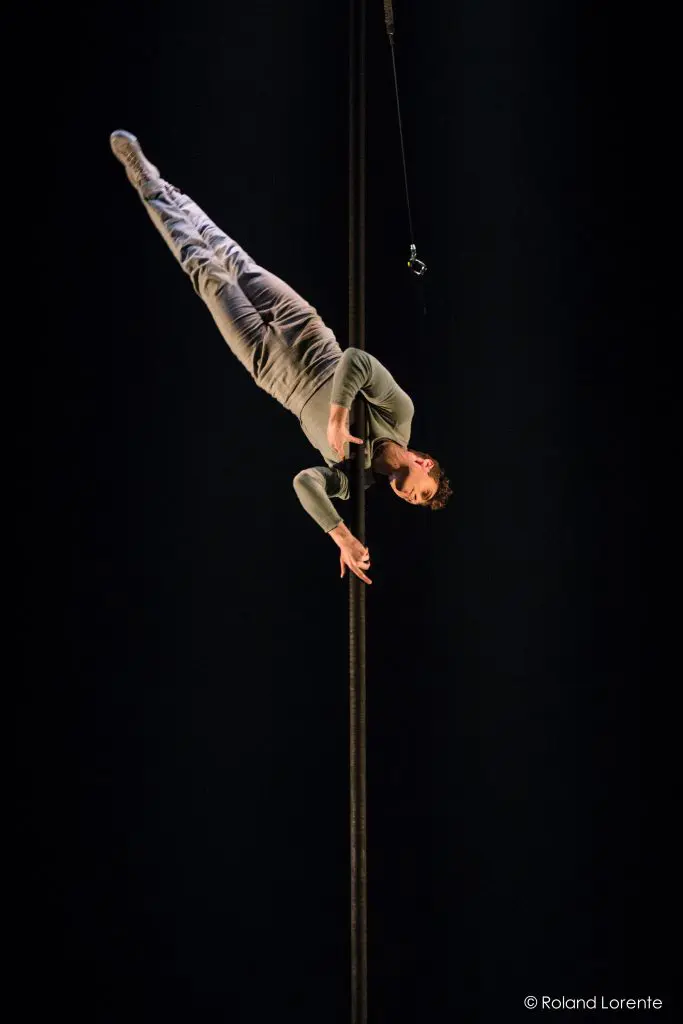 Un artiste vêtu de gris est suspendu à l'envers, agrippant un poteau vertical avec un bras et une jambe sur un fond sombre. Un câble est attaché à la perche au-dessus, mettant en valeur les remarquables talents d'artiste aérien de Ron Oppenheimer.