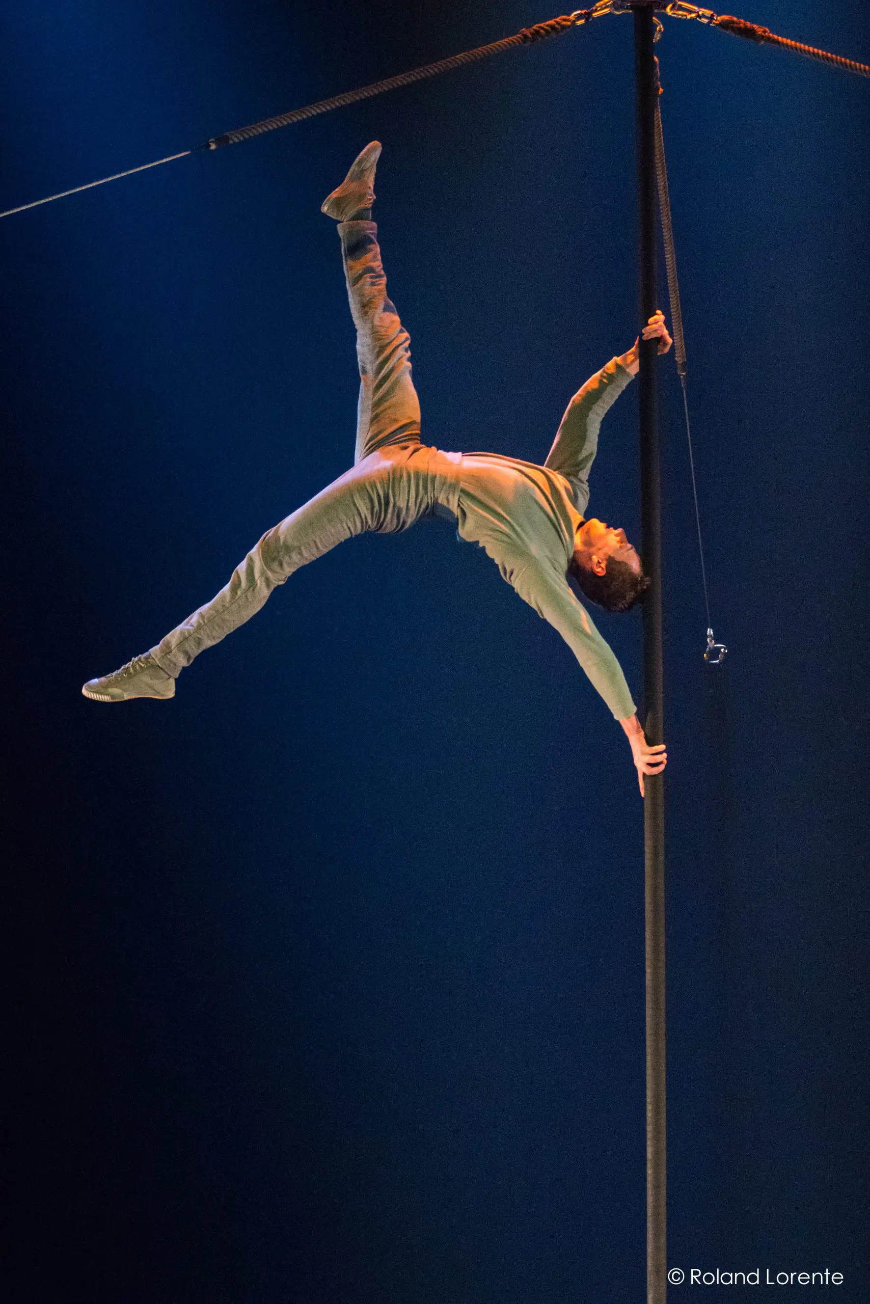 Ron Oppenheimer, vêtu de vert, est suspendu horizontalement à un grand poteau vertical, qu'il saisit d'une main et d'un pied, tandis que l'autre bras et l'autre jambe s'étendent vers l'extérieur, sur un fond éclairé en bleu foncé.