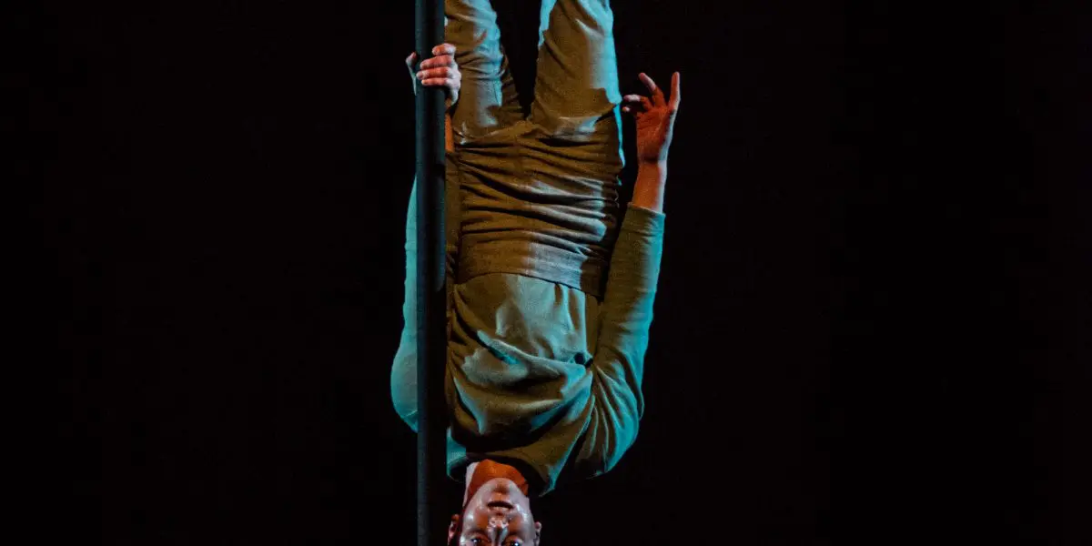 Un artiste, Ron Oppenheimer, est suspendu à l'envers à une corde verticale dans un espace sombre, vêtu de vêtements décontractés et de baskets, éclairé par un projecteur. Crédit photo : Roland Lorente.