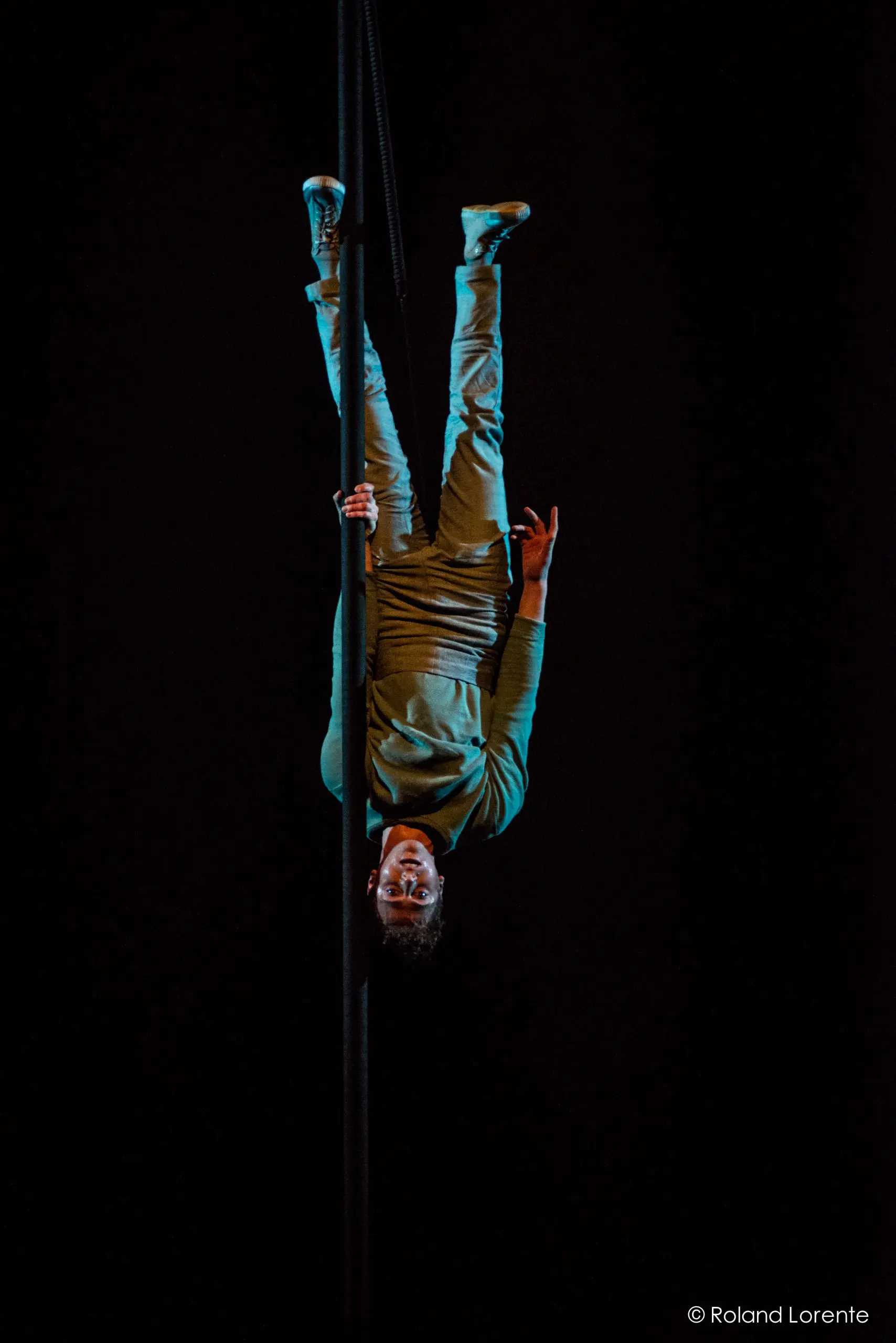 Un artiste, Ron Oppenheimer, est suspendu à l'envers à une corde verticale dans un espace sombre, vêtu de vêtements décontractés et de baskets, éclairé par un projecteur. Crédit photo : Roland Lorente.
