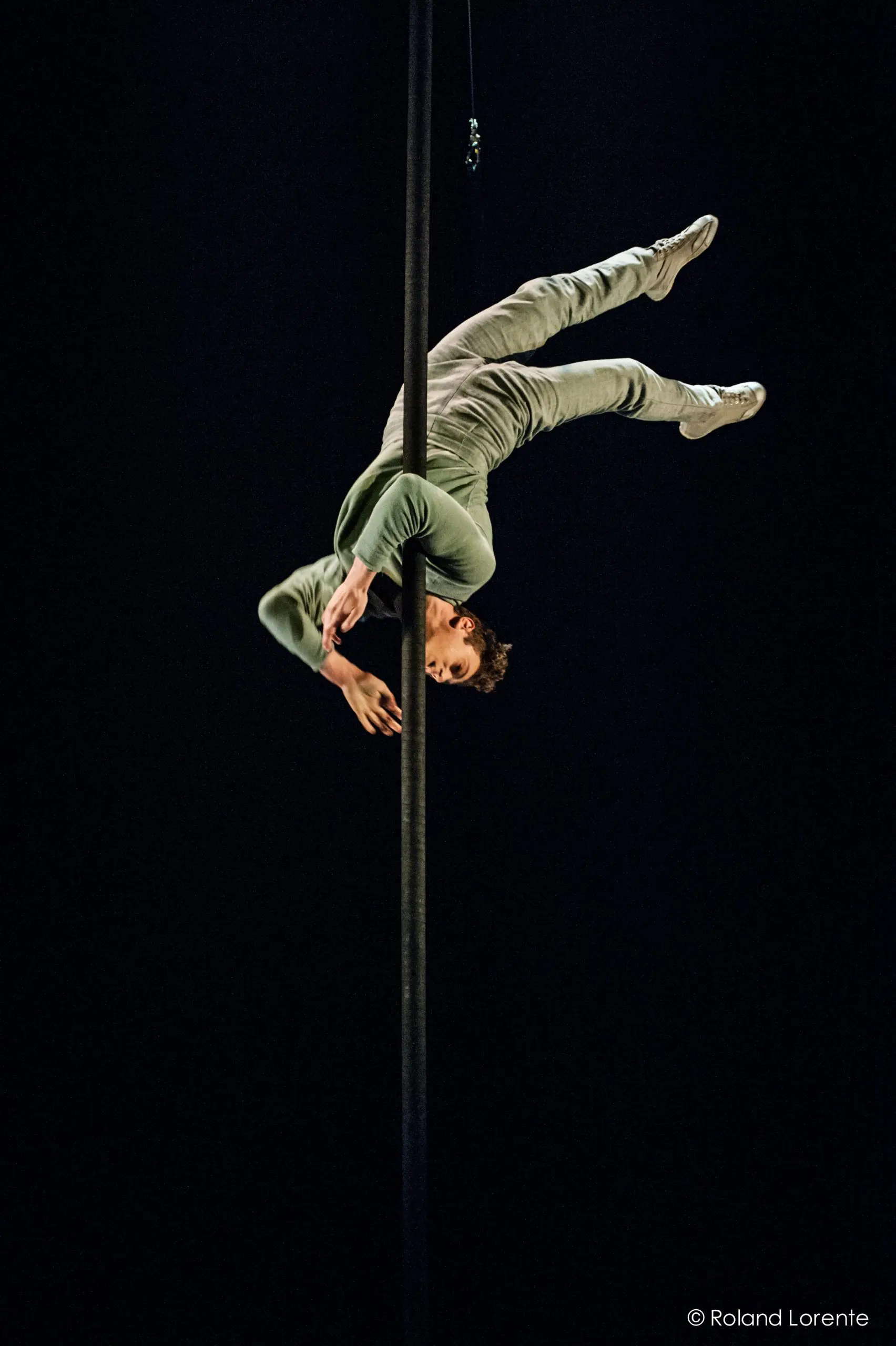 Un acrobate vêtu d'un costume vert clair prend la pose à l'envers sur un poteau vertical sur un fond sombre, mettant en valeur la force et l'équilibre dans le style caractéristique de Ron Oppenheimer.