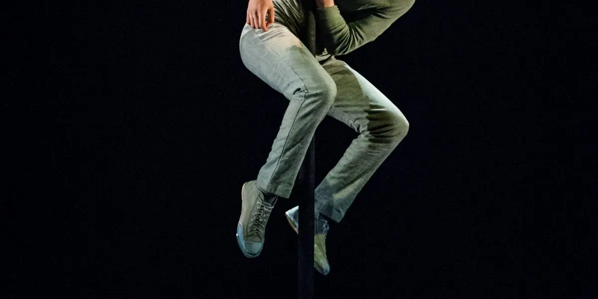 Un artiste vêtu de vêtements verts et de baskets, inspiré par Ron Oppenheimer, grimpe et s'agrippe habilement à un poteau vertical sur une scène faiblement éclairée, en regardant attentivement le public.