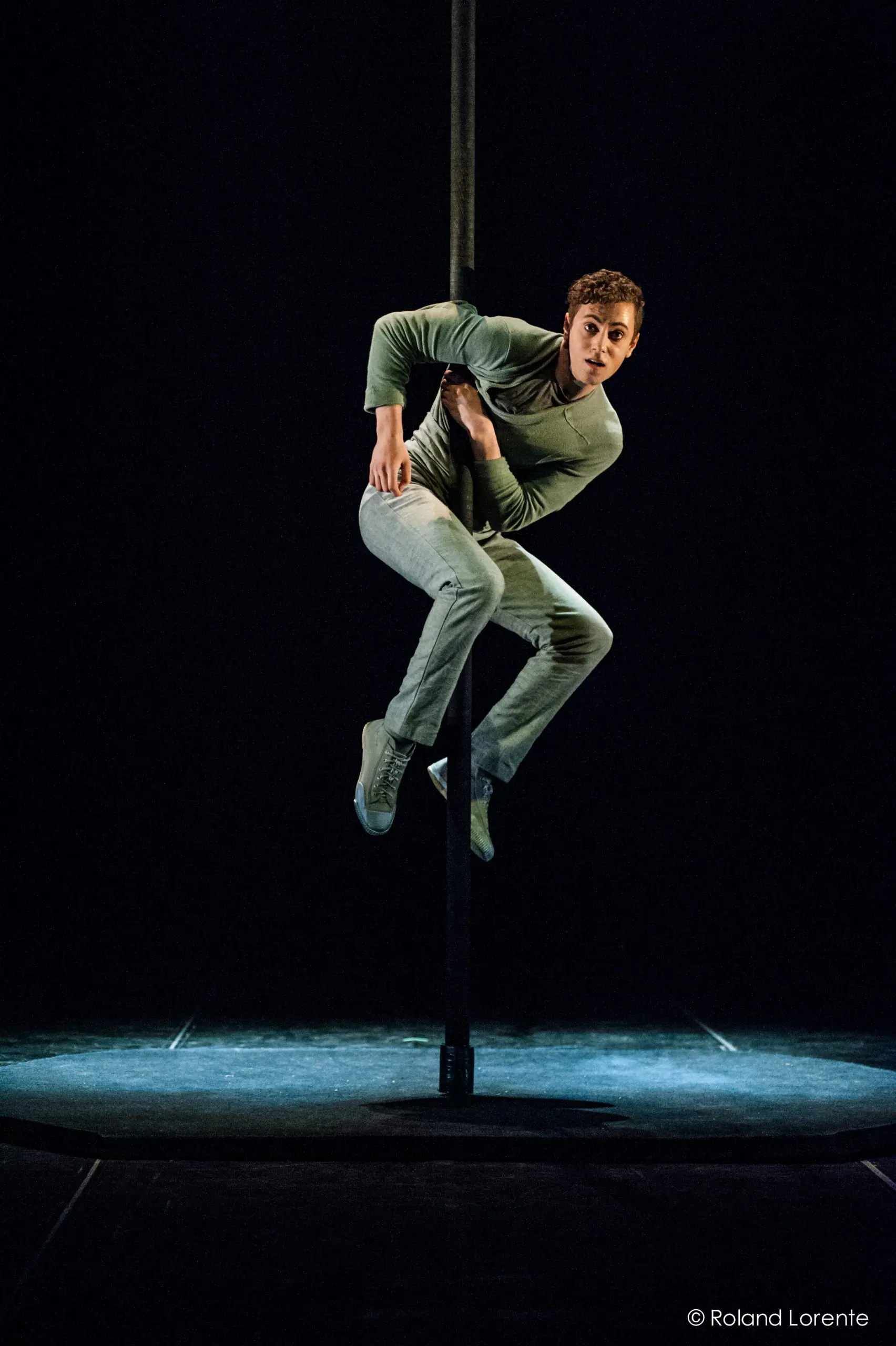Un artiste vêtu de vêtements verts et de baskets, inspiré par Ron Oppenheimer, grimpe et s'agrippe habilement à un poteau vertical sur une scène faiblement éclairée, en regardant attentivement le public.