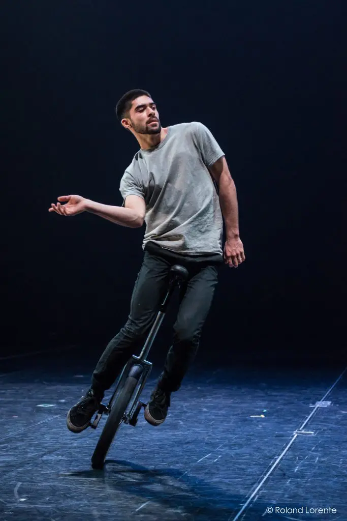 Un homme vêtu d'un t-shirt gris et d'un pantalon noir se tient en équilibre sur un monocycle sur une scène sombre, penché d'un côté, un bras tendu et l'autre plié, comme si Ruben Ingwersen était en train d'exécuter une danse ou un tour.