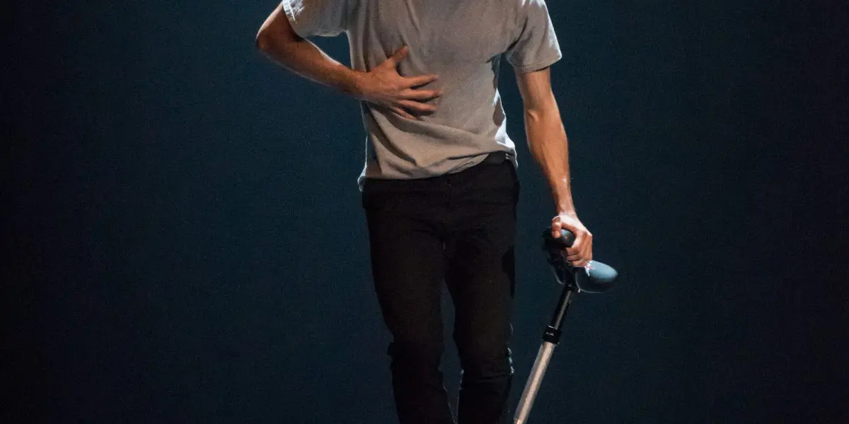 Ruben Ingwersen, vêtu d'un t-shirt gris et d'un pantalon noir, roule sur un monocycle sur une scène sombre et éclairée, une main sur la poitrine et l'autre tenant le siège du monocycle.