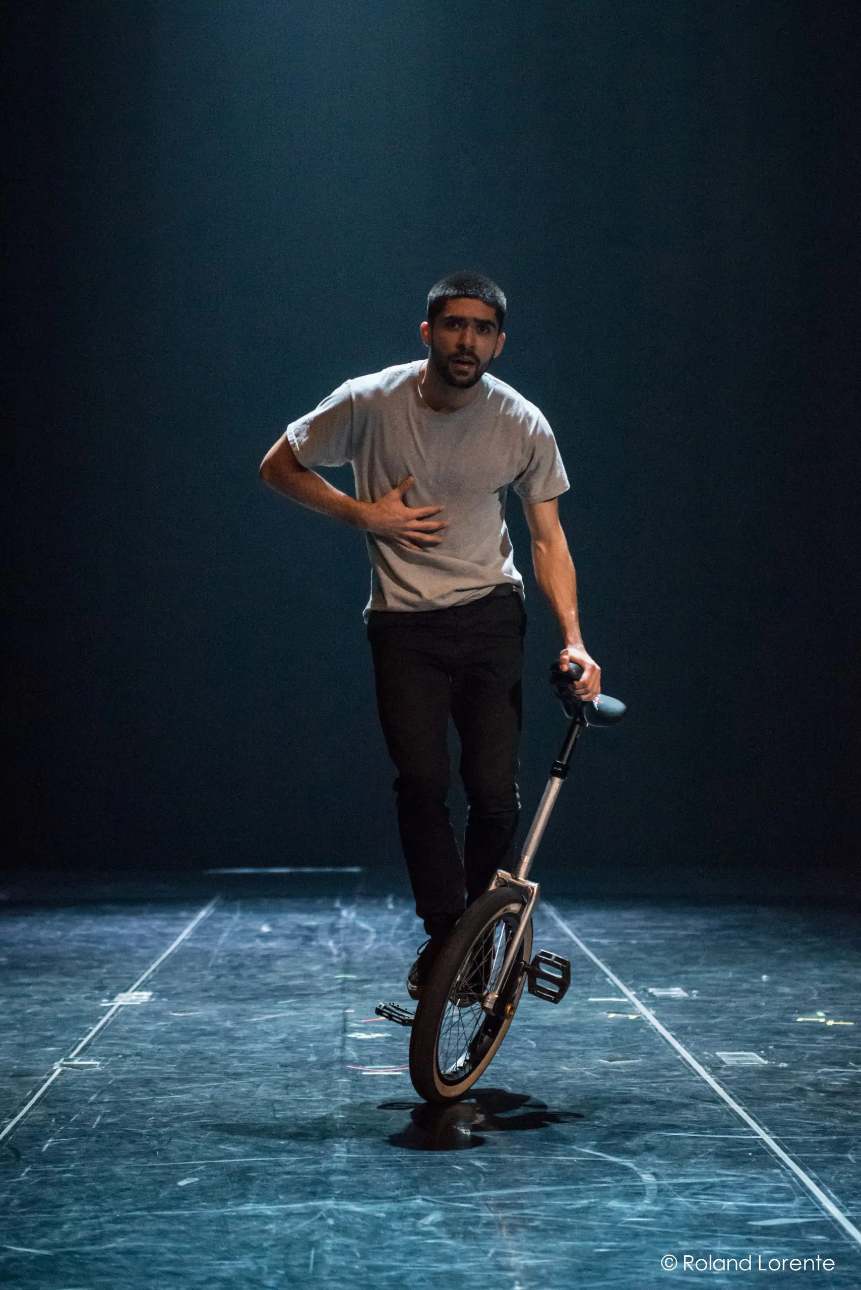 Ruben Ingwersen, vêtu d'un t-shirt gris et d'un pantalon noir, roule sur un monocycle sur une scène sombre et éclairée, une main sur la poitrine et l'autre tenant le siège du monocycle.