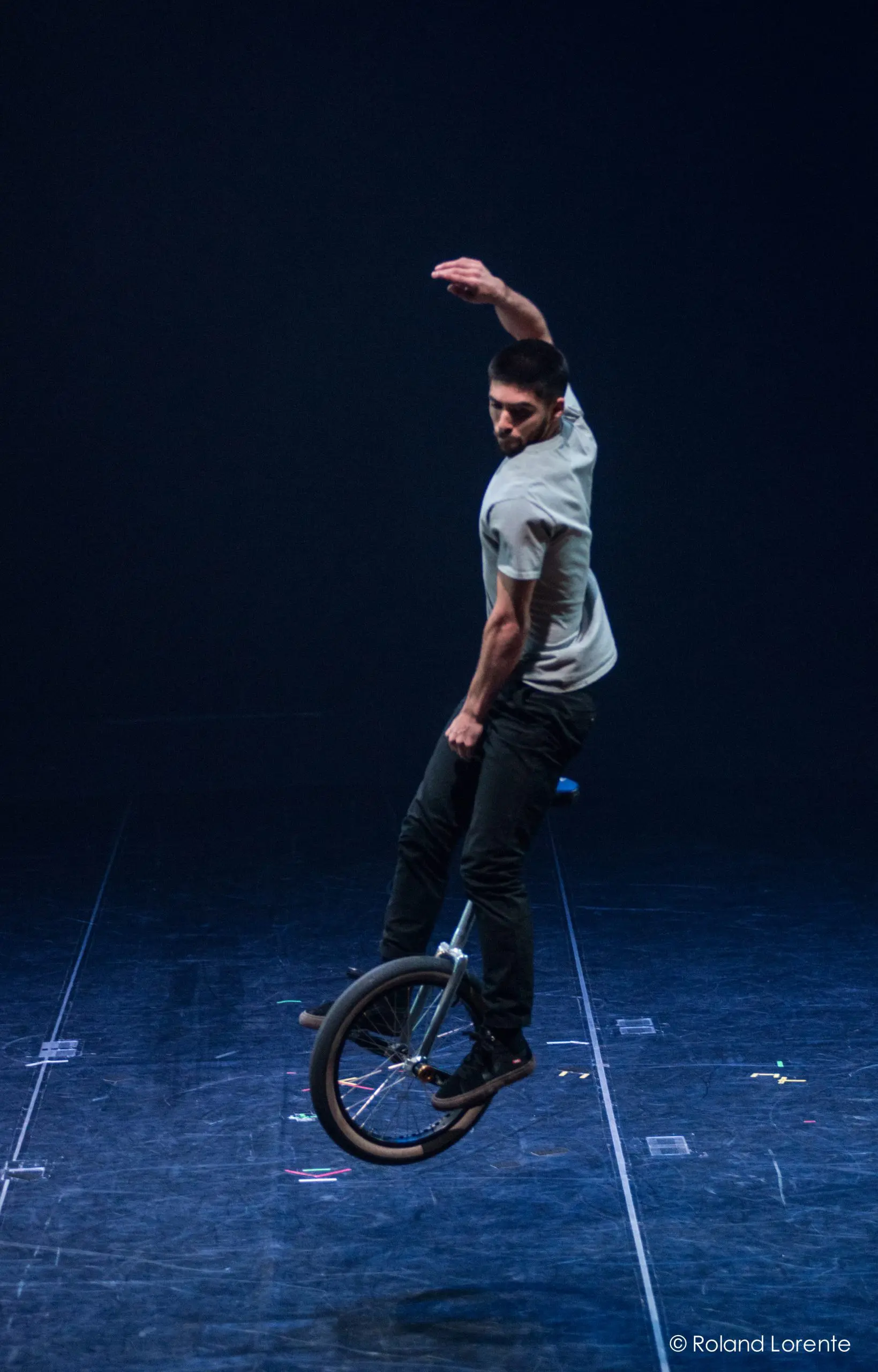 Ruben Ingwersen se tient en équilibre sur un monocycle sur une scène sombre, penché d'un côté, un bras levé et l'autre baissé, semblant exécuter un tour sous un éclairage dramatique.
