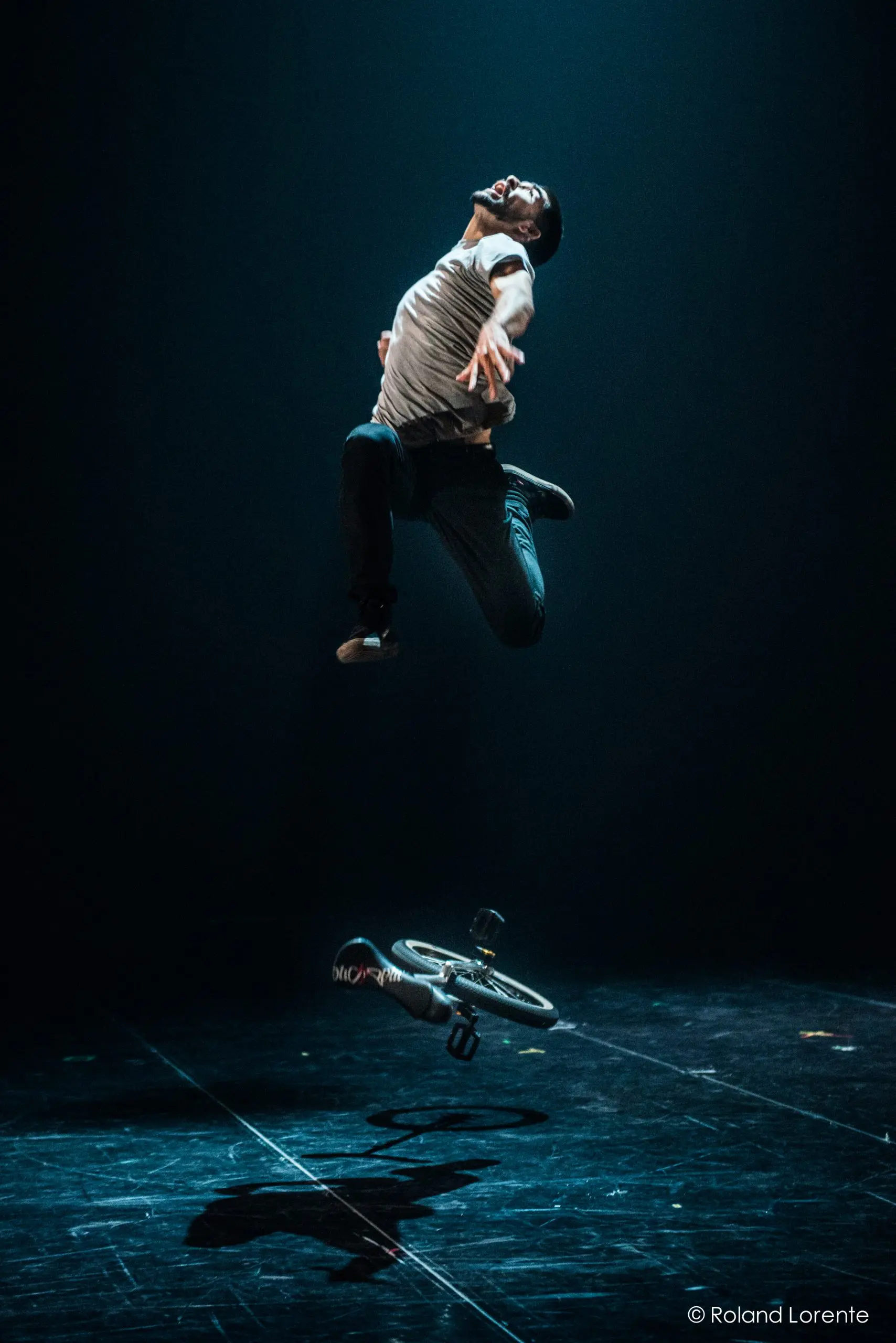 Une personne saute spectaculairement en l'air au-dessus d'un petit vélo, éclairé par un projecteur sur une scène sombre. Rappelant le style caractéristique de Ruben Ingwersen, le vélo semble être en mouvement et projette une ombre sur le sol.