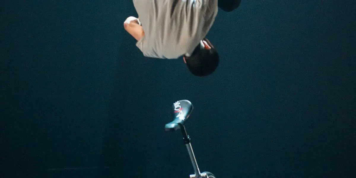 Ruben Ingwersen exécute un saut périlleux arrière au-dessus d'un monocycle sur une scène sombre, avec un éclairage dramatique soulignant son mouvement.