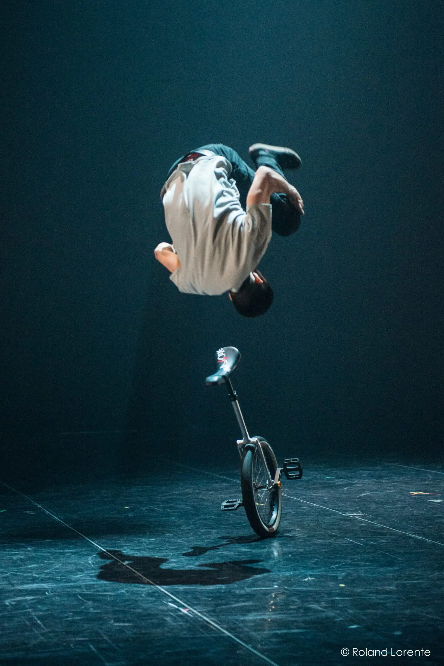 Ruben Ingwersen exécute un saut périlleux arrière au-dessus d'un monocycle sur une scène sombre, avec un éclairage dramatique soulignant son mouvement.