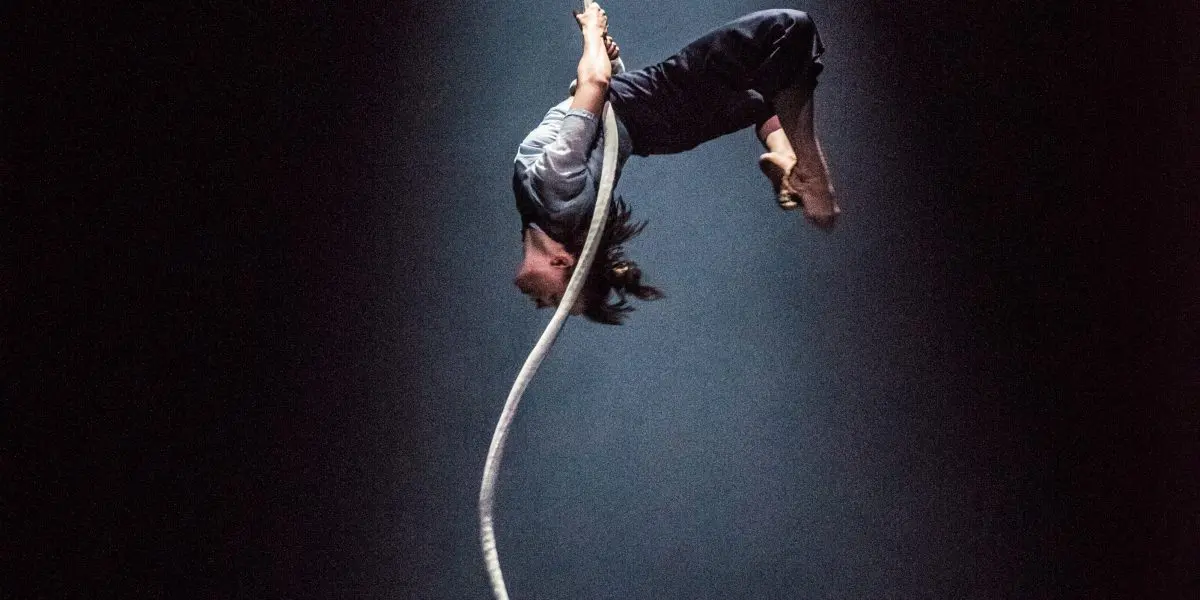 Sandy Tugwood, artiste aérienne, est suspendue à l'envers à une corde blanche sous un projecteur, avec des bâches en plastique sur le sol sombre de la scène.