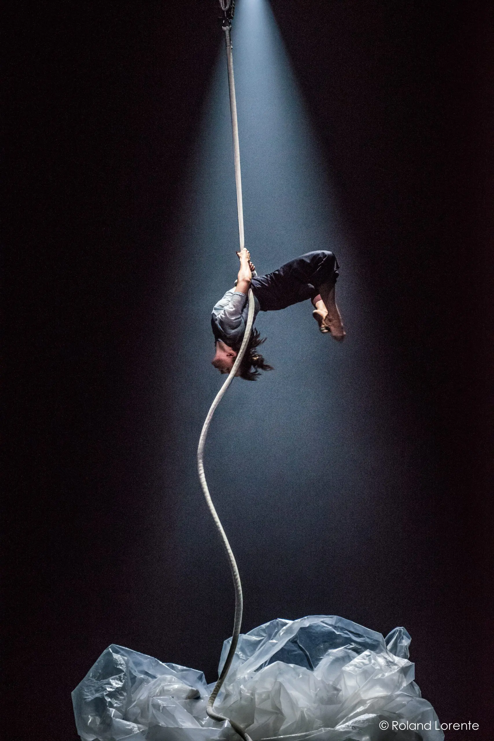 Sandy Tugwood, artiste aérienne, est suspendue à l'envers à une corde blanche sous un projecteur, avec des bâches en plastique sur le sol sombre de la scène.