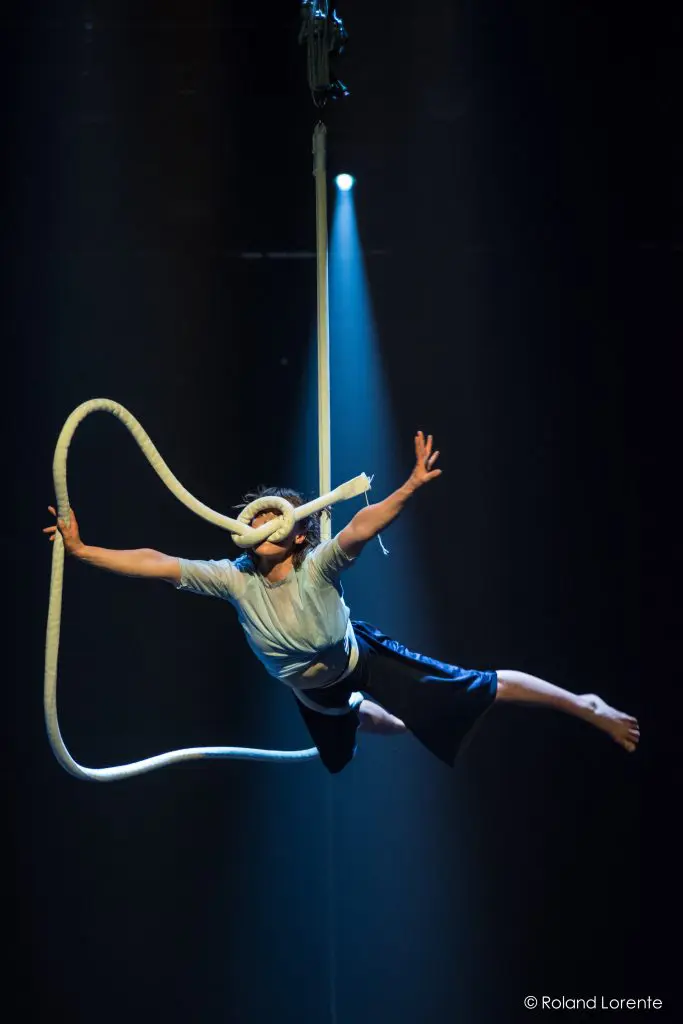 Sandy Tugwood, une artiste vêtue d'une chemise bleu clair et d'un short sombre, est suspendue à une corde aérienne blanche, bras et jambes gracieusement tendus, éclairée par un projecteur sur une scène sombre.