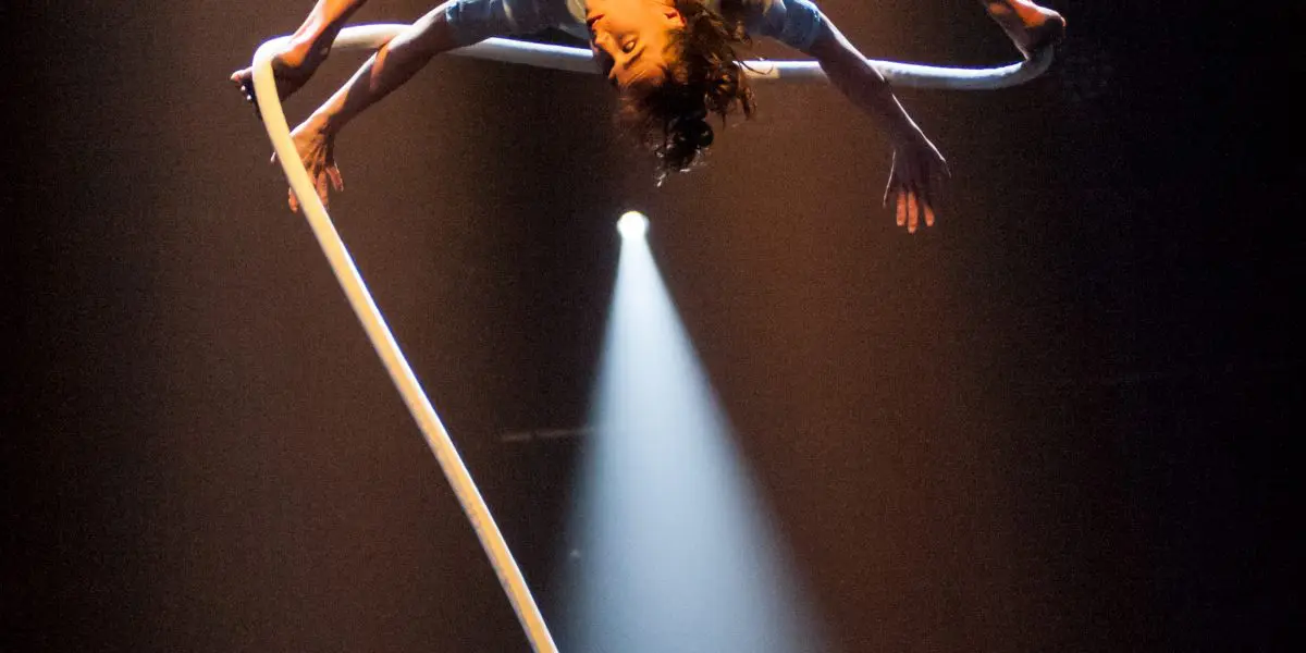 Une artiste aérienne, Sandy Tugwood, est suspendue la tête en bas, en équilibre sur une corde blanche. Elle étire gracieusement ses membres sous un éclairage dramatique, un projecteur l'éclairant sur un fond sombre.