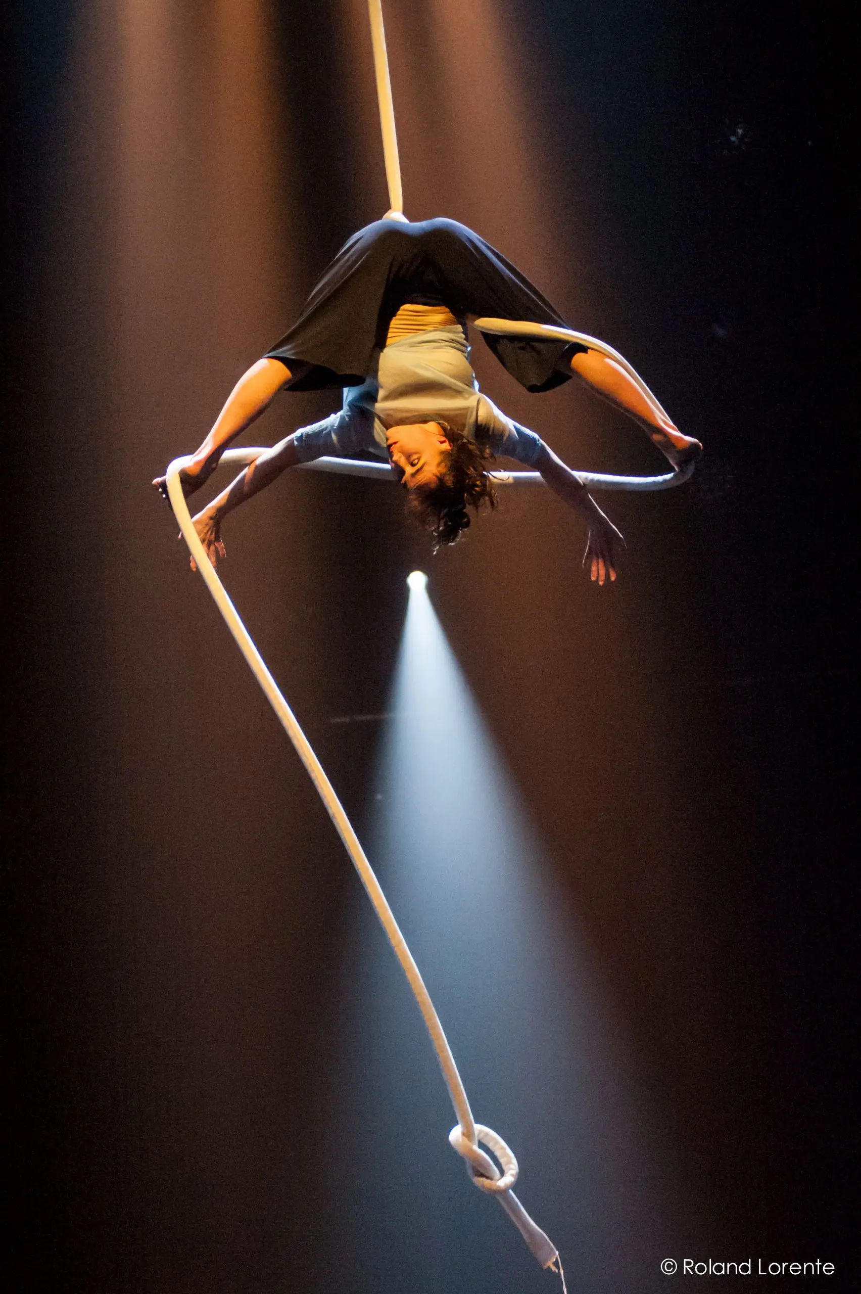 Une artiste aérienne, Sandy Tugwood, est suspendue la tête en bas, en équilibre sur une corde blanche. Elle étire gracieusement ses membres sous un éclairage dramatique, un projecteur l'éclairant sur un fond sombre.