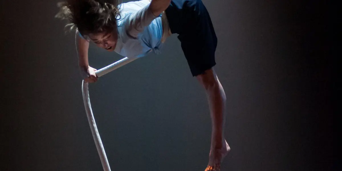 Une personne aux cheveux longs et aux pieds nus est suspendue la tête en bas, exécutant un numéro d'acrobatie aérienne avec une corde blanche sur un fond sombre et éclairé - une scène captivante qui rappelle le style caractéristique de Sandy Tugwood.