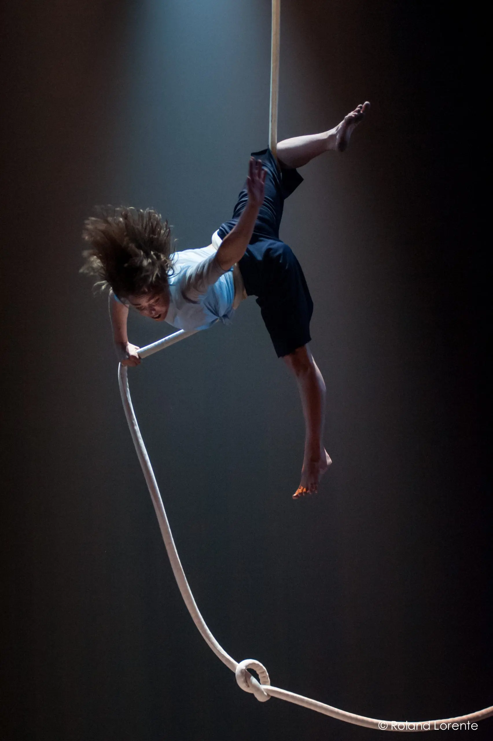Une personne aux cheveux longs et aux pieds nus est suspendue la tête en bas, exécutant un numéro d'acrobatie aérienne avec une corde blanche sur un fond sombre et éclairé - une scène captivante qui rappelle le style caractéristique de Sandy Tugwood.