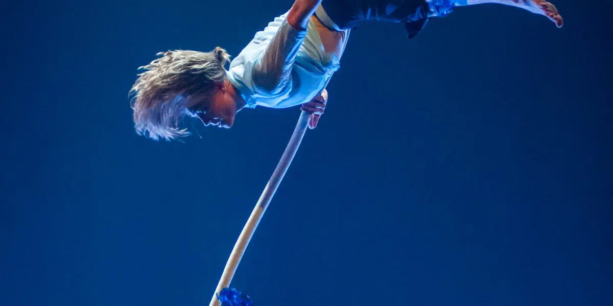 Un enfant effectue un numéro d'acrobatie aérienne, suspendu à l'envers à une corde sur un fond bleu, avec des papillons bleus éparpillés autour - une scène fantaisiste qui rappelle les mondes enchanteurs de Sandy Tugwood.