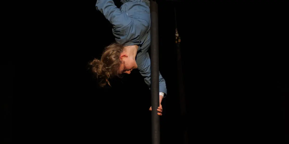 L'artiste de cirque Sara McDonald, vêtue de bleu, se tient en équilibre à l'envers sur un poteau vertical, les jambes pliées et agrippées au poteau, sur un fond sombre.