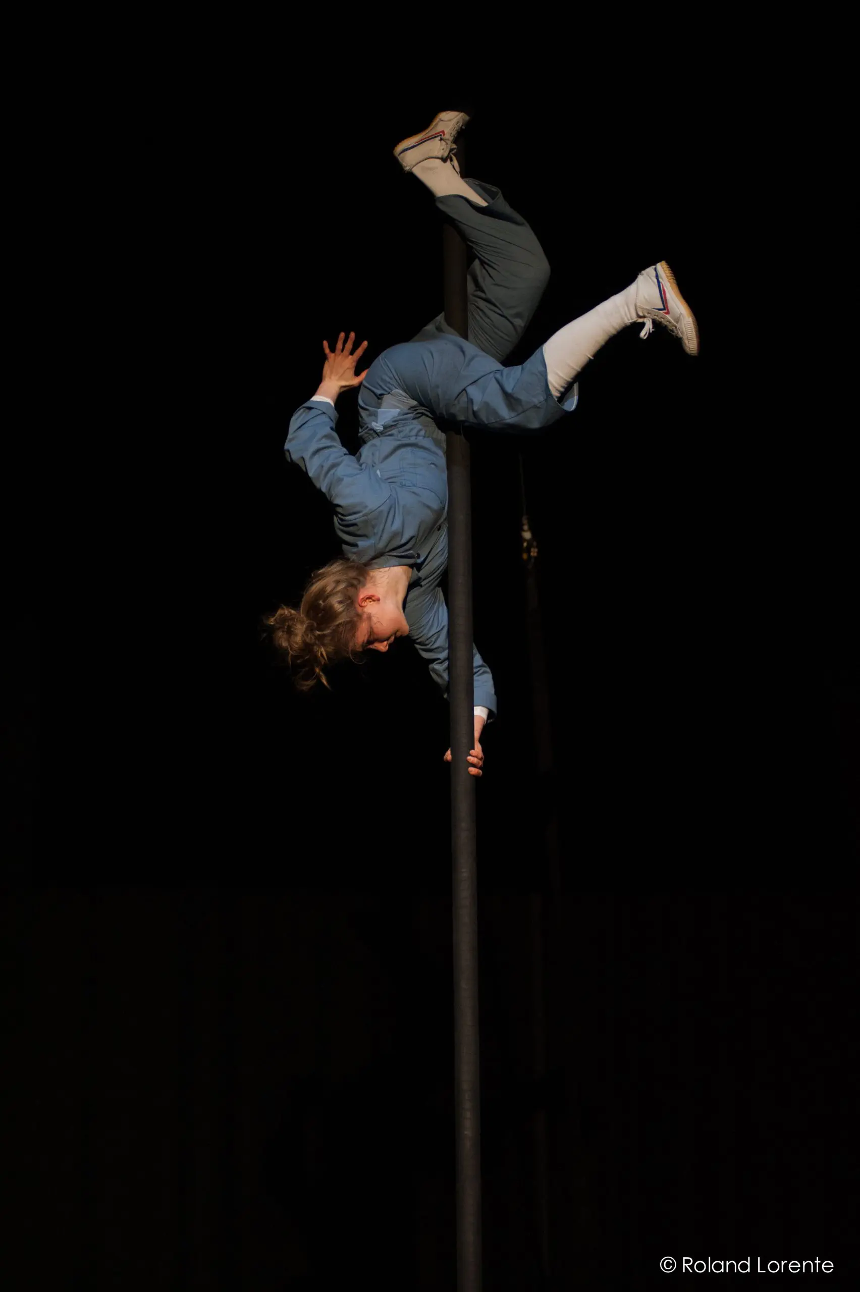 L'artiste de cirque Sara McDonald, vêtue de bleu, se tient en équilibre à l'envers sur un poteau vertical, les jambes pliées et agrippées au poteau, sur un fond sombre.