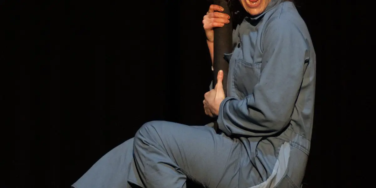 Sara McDonald, vêtue d'une combinaison bleue et de baskets blanches, s'accroche à un poteau vertical, la bouche grande ouverte et les yeux remplis de stupeur sur un fond sombre.