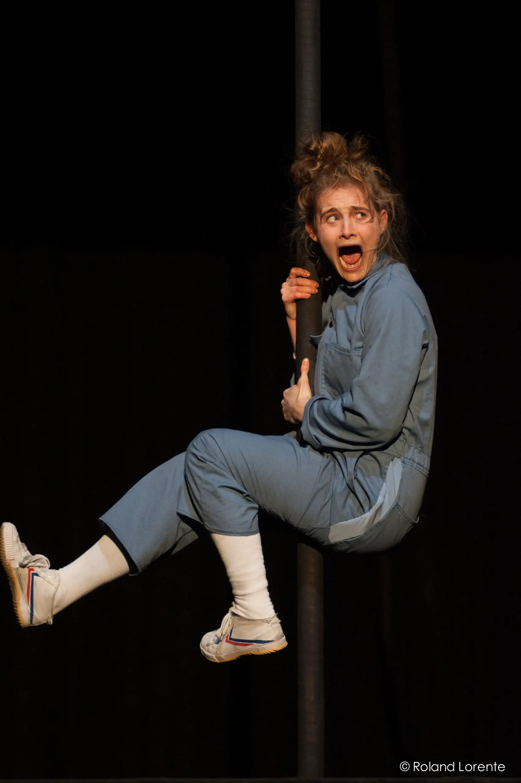 Sara McDonald, vêtue d'une combinaison bleue et de baskets blanches, s'accroche à un poteau vertical, la bouche grande ouverte et les yeux remplis de stupeur sur un fond sombre.