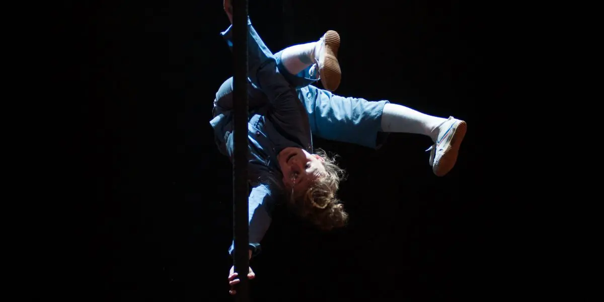 Un artiste vêtu de vêtements bleus et de chaussures blanches est suspendu à un poteau vertical sur une scène sombre, avec un éclairage dramatique qui met en valeur sa silhouette - un acte qui rappelle les performances captivantes de Sara McDonald.