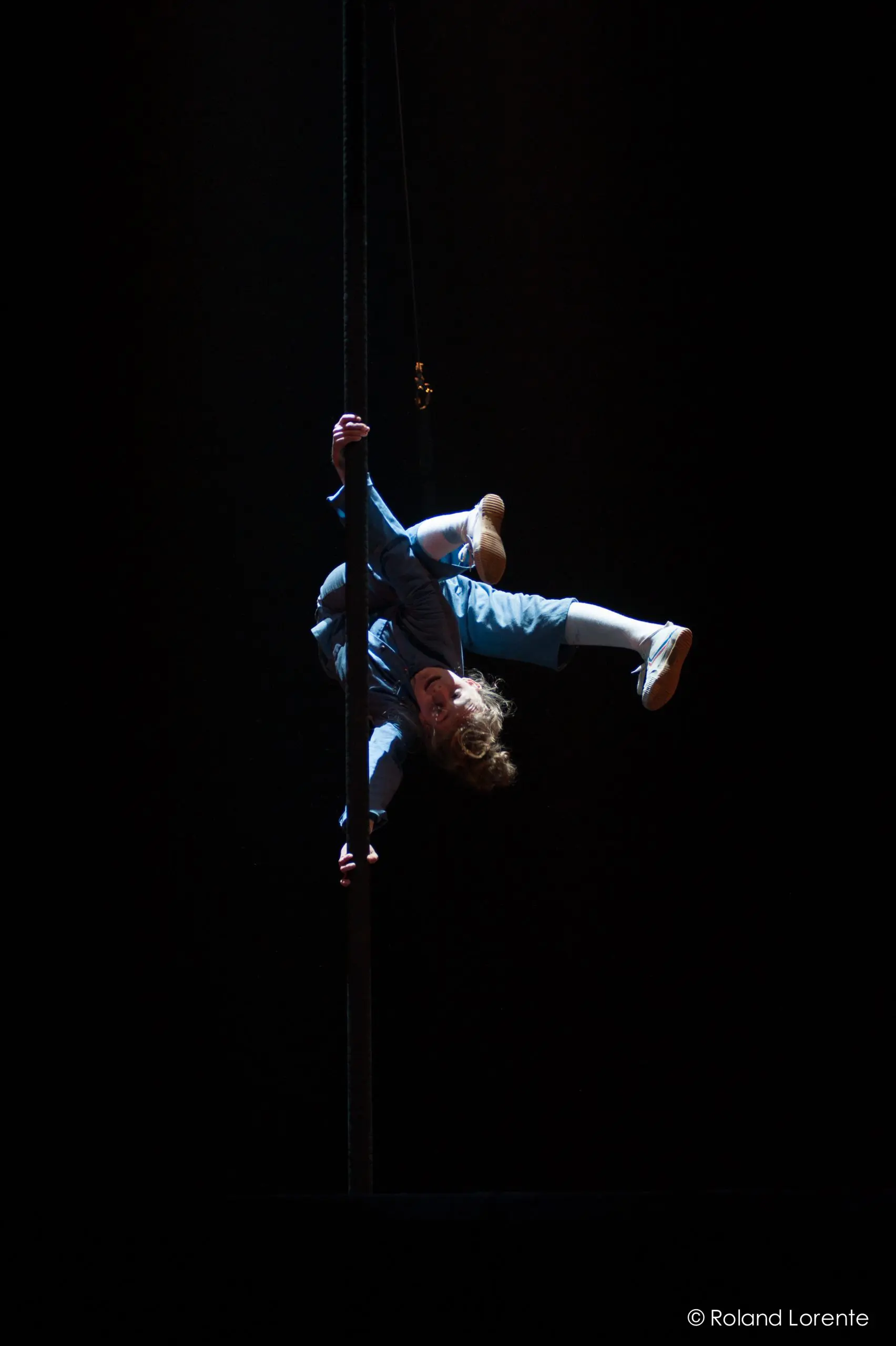 Un artiste vêtu de vêtements bleus et de chaussures blanches est suspendu à un poteau vertical sur une scène sombre, avec un éclairage dramatique qui met en valeur sa silhouette - un acte qui rappelle les performances captivantes de Sara McDonald.