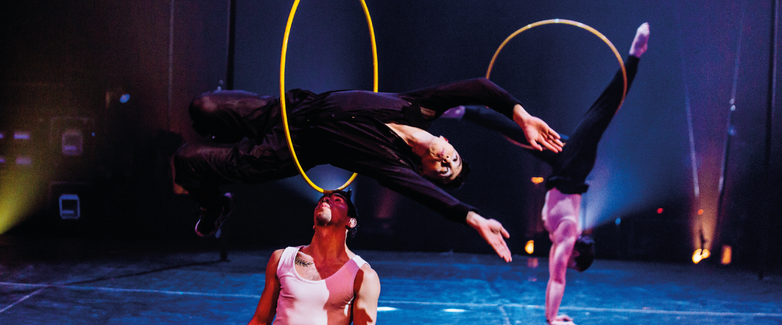 Deux acrobates se produisent sur scène : l'un saute à travers un cerceau jaune tenu par un autre, tous deux dans des poses dynamiques. Un troisième artiste fait un poirier sous un éclairage dramatique, vous invitant à découvrir le monde vibrant du mouvement de l'École.