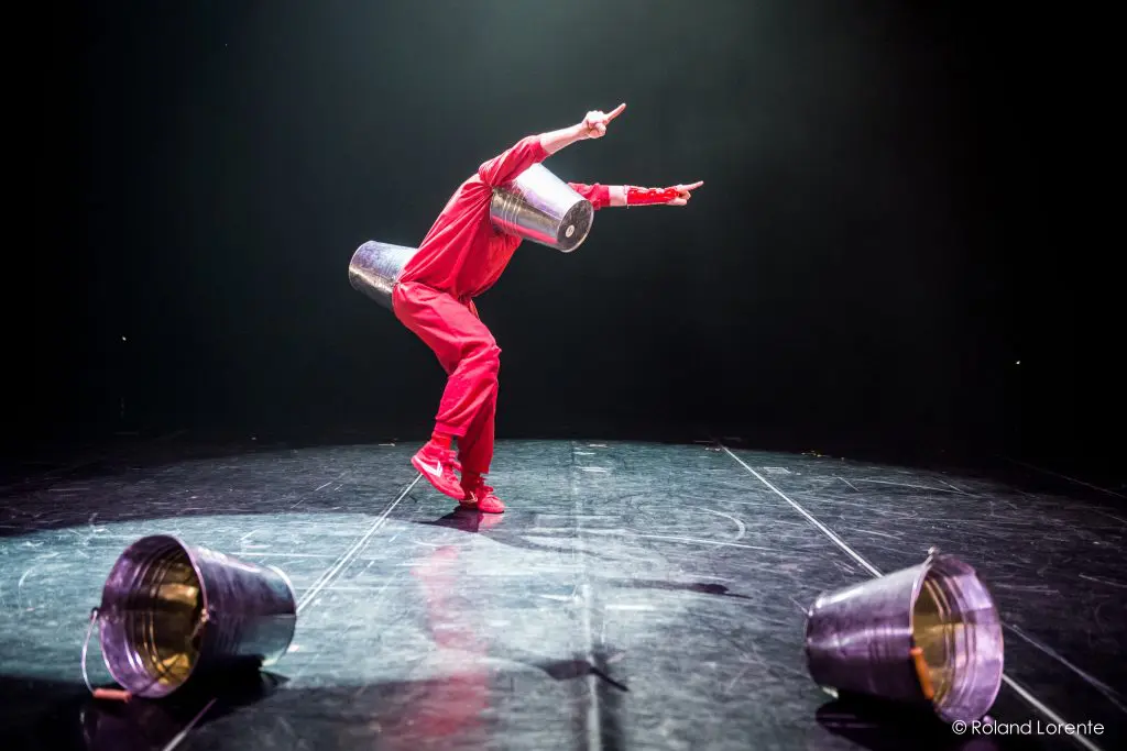 Éline Guélat, vêtue d'une tenue rouge, se tient en équilibre sur une jambe, les bras tendus, des poubelles métalliques sur la tête et le bras. Deux autres poubelles gisent sur le sol de la scène faiblement éclairée, ajoutant à l'intrigue de la scène.