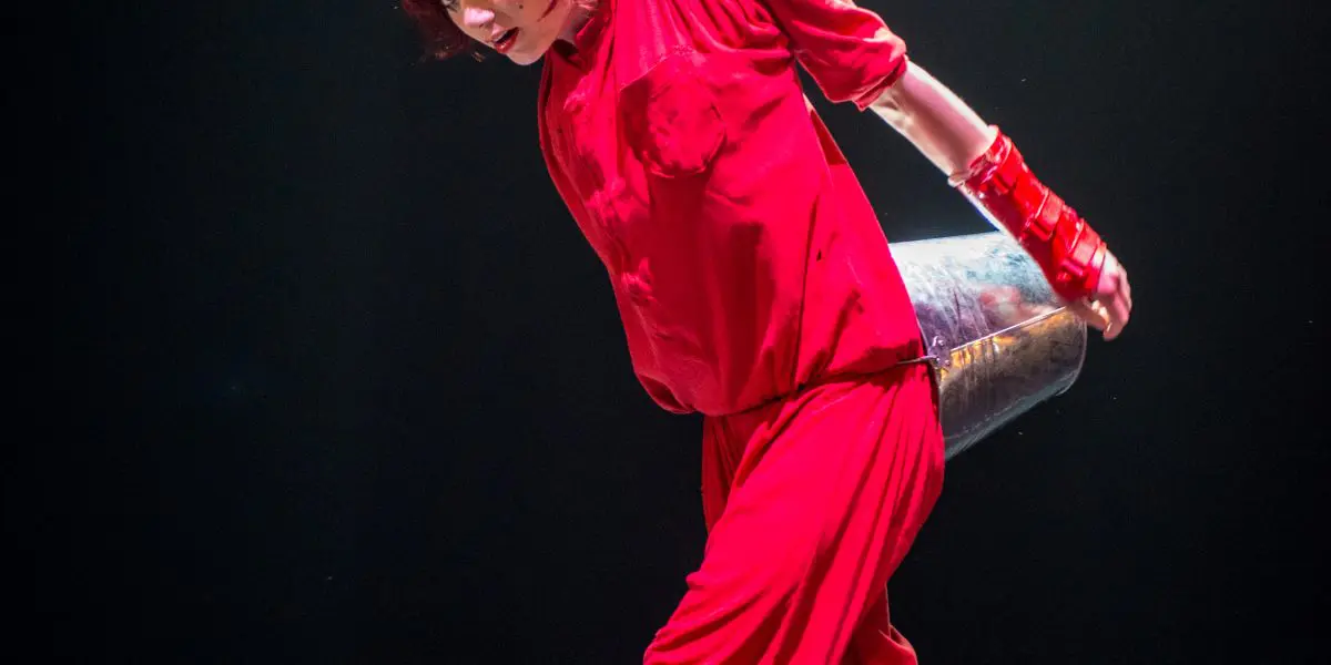 Éline Guélat, vêtue de rouge, le visage peint et les gants assortis, prend une pose ludique et dramatique sur une scène sombre - elle tient en équilibre un seau métallique sur son dos et se tient debout, un pied à l'intérieur d'un autre seau.