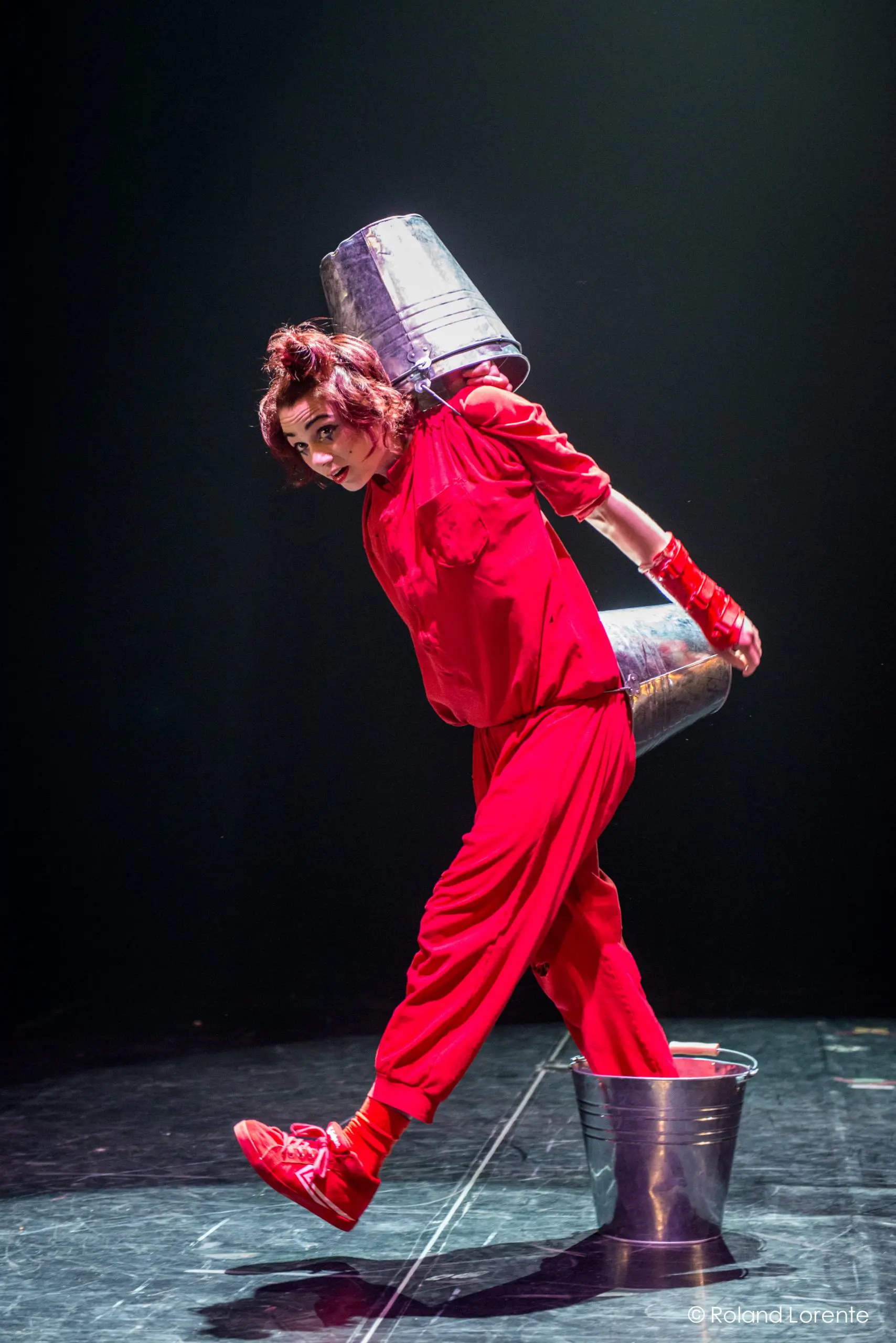 Éline Guélat, vêtue de rouge, le visage peint et les gants assortis, prend une pose ludique et dramatique sur une scène sombre - elle tient en équilibre un seau métallique sur son dos et se tient debout, un pied à l'intérieur d'un autre seau.