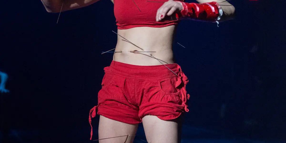 Un interprète vêtu d'un crop top rouge et d'un short se tient sur scène, tenant de fines tiges ou des fils, avec d'autres tiges attachées à leurs membres, évoquant un thème de marionnette sous un éclairage bleu dramatique - une pièce évocatrice d'Éline Guélat.