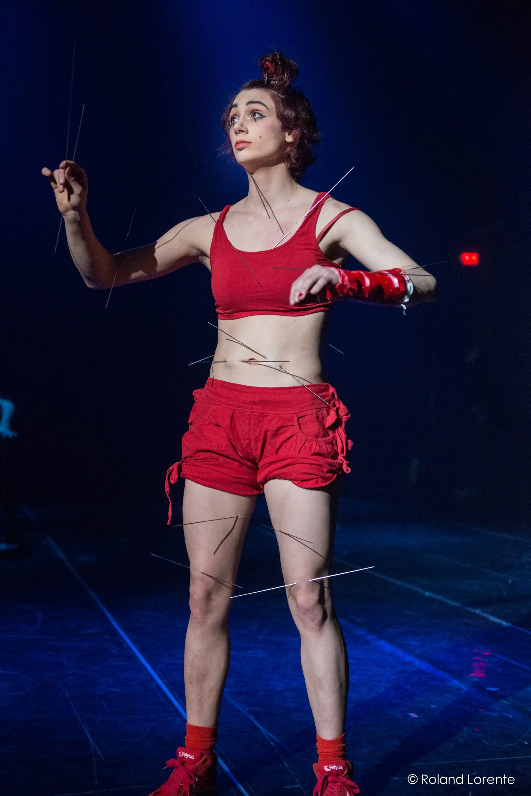 Un interprète vêtu d'un crop top rouge et d'un short se tient sur scène, tenant de fines tiges ou des fils, avec d'autres tiges attachées à leurs membres, évoquant un thème de marionnette sous un éclairage bleu dramatique - une pièce évocatrice d'Éline Guélat.